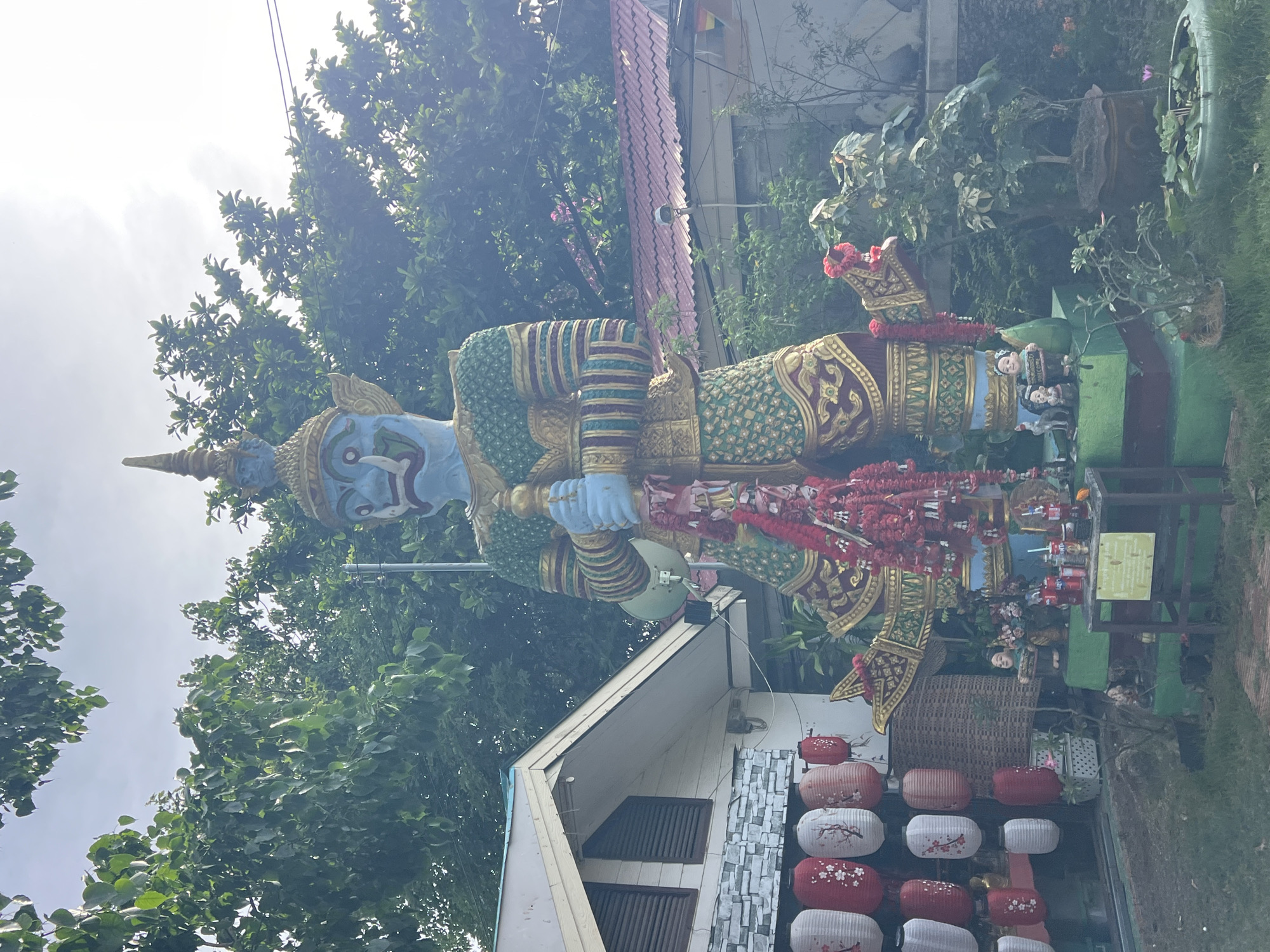 The image depicts a large, blue-skinned statue of a mythological figure, likely a giant or a demon, standing in front of a building. The statue is elaborately decorated with green and gold patterned clothing, armbands, and a headdress. It holds a golden staff. Red garlands hang from the statue's chest and arms.
To the left of the statue, a building with a white, sloped roof and horizontal blinds on its windows is visible. A wall with stone-like cladding is below the windows. Several paper lanterns, red and white with floral designs, are hanging from the building's exterior.
In front of the statue, a tiered platform painted green and red serves as a base. On this platform, there are small figurines, including two children holding a sign that says "WELCOME," and various offerings such as bottles of drinks and what appear to be incense sticks. A small table with a yellow sign is placed in front of the platform.
Behind the statue and to the right, parts of a red-tiled roof and more greenery are visible, suggesting an outdoor setting. The environment appears to be a courtyard or a garden area. The overall impression is that this is a religious or cultural site.
The location is identified as Baan Plai Laem, Thailand. The scene is set outdoors, with trees and foliage framing the statue and building. The sky is overcast or bright with diffused light.