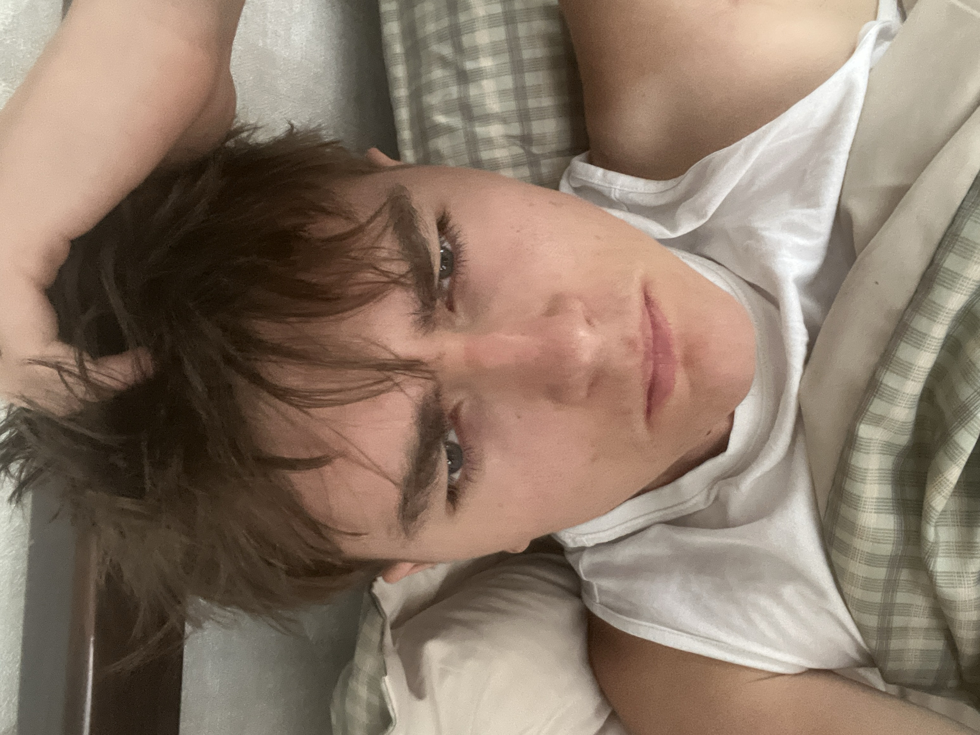 A young person with messy brown hair and a white tank top is lying in bed. Their hand is resting on their head, and their face is turned towards the camera with a slightly furrowed brow. The background shows pillows and a plaid blanket, suggesting a bedroom setting. The image does not provide any information about the city of Sobishchytsi, Ukraine.