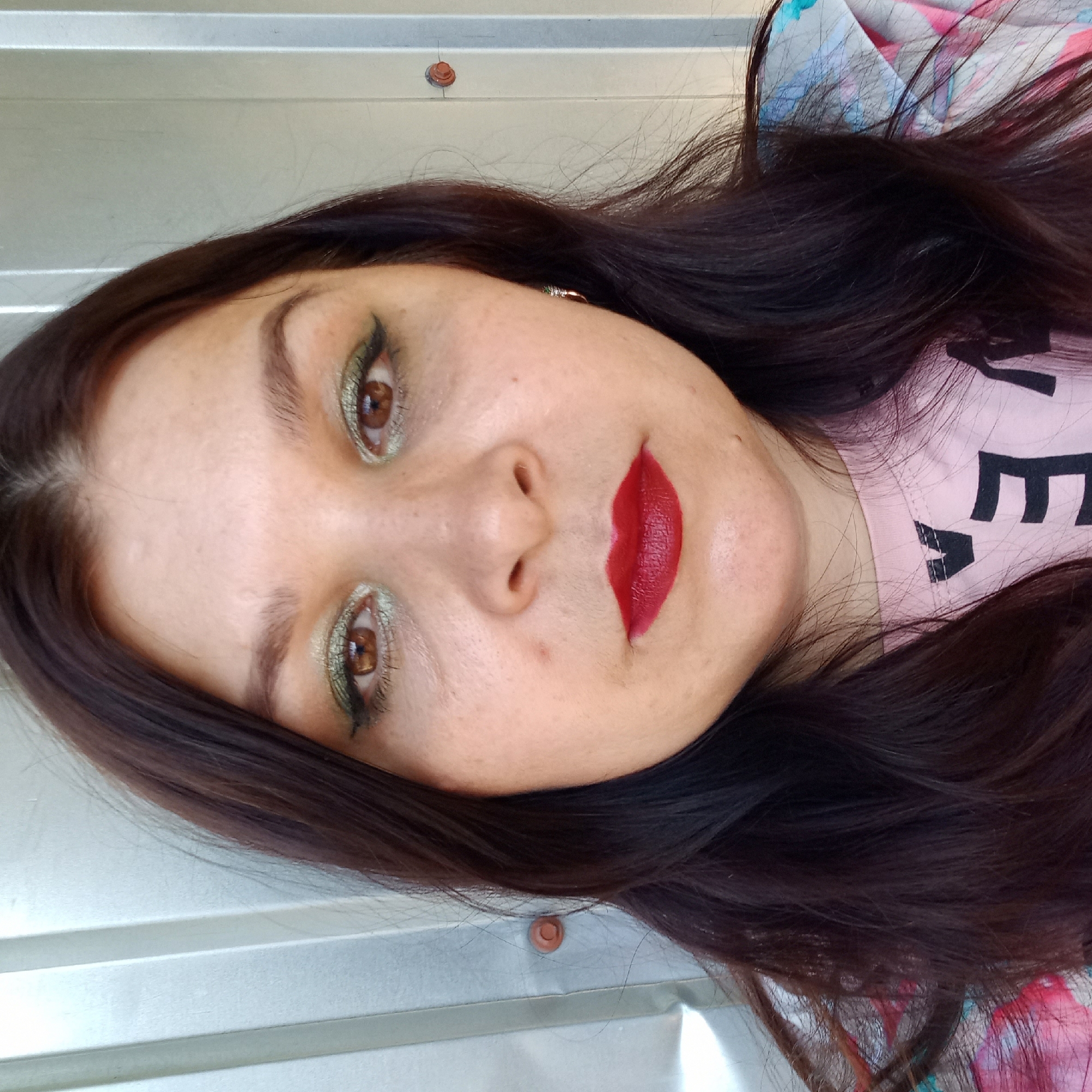 A medium shot, head-and-shoulders, selfie of a woman with long, dark brown hair, brown eyes, and fair skin. She is wearing a pink t-shirt and a colorful shirt with blue, pink, and white abstract patterns. Her makeup includes green eyeshadow and dark red lipstick. The background is a corrugated metal wall. The woman is indoors or outdoors with natural light. There are no visible landmarks or details to indicate a specific location in Kropyvnytskyi, Ukraine.