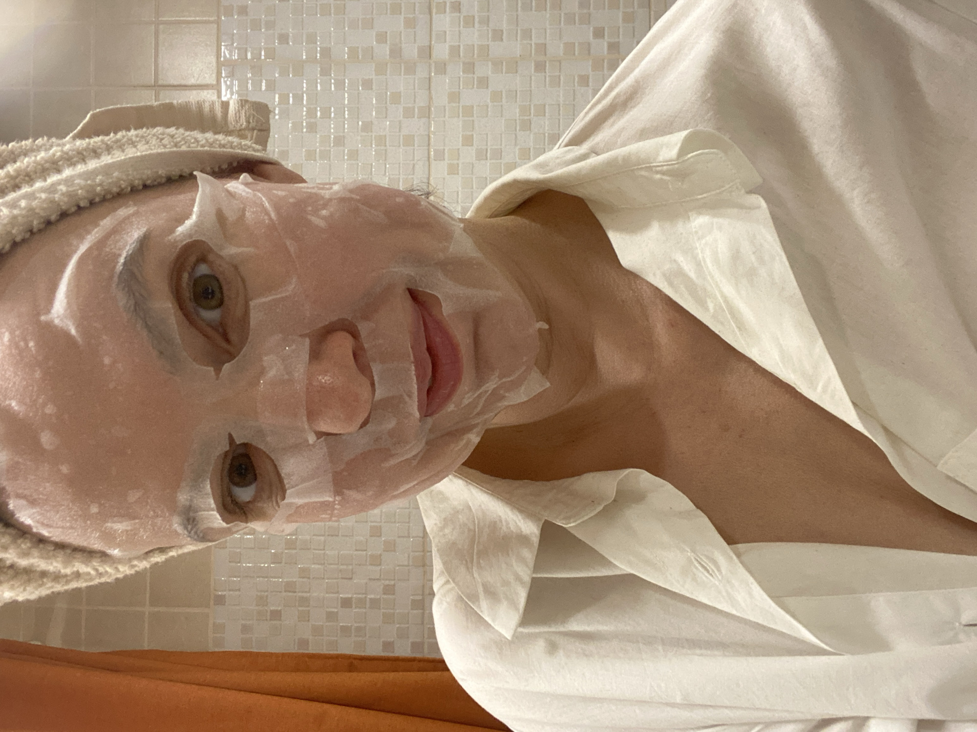 The image depicts a person indoors, wearing a white sheet mask and a towel wrapped around their head. The person is also wearing a white collared shirt. The background shows tiled walls, possibly in a bathroom setting. No information suggests the location to be Chaikovsky, Russia.