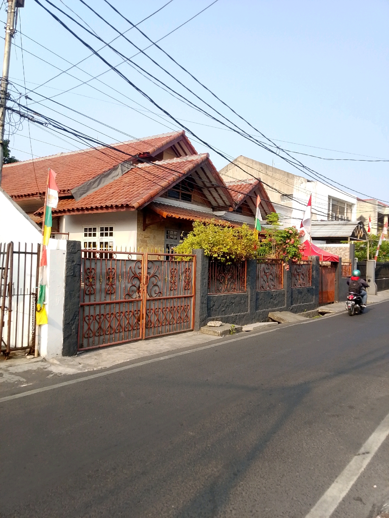 The image captures a street scene in East Jakarta, Indonesia, on a clear day under a pale blue sky. Overhead, a dense network of power lines crisscrosses the frame. In the mid-ground, a row of residential buildings lines the street, characterized by their terracotta tiled roofs and decorative metal gates. Several Indonesian flags are visible, attached to poles and flags planted in front of the houses, suggesting a national celebration or commemoration. The buildings exhibit a mix of architectural styles, with some featuring stone accents and others smooth, light-colored facades. A motorcyclist wearing a helmet and a dark jacket is seen riding away from the camera on the asphalt road that occupies the foreground and extends into the distance. The road shows a white line marking the lane. Lush green foliage and flowering plants are present in front of some of the houses, softening the urban landscape. The ground features a concrete sidewalk and a drainage ditch.