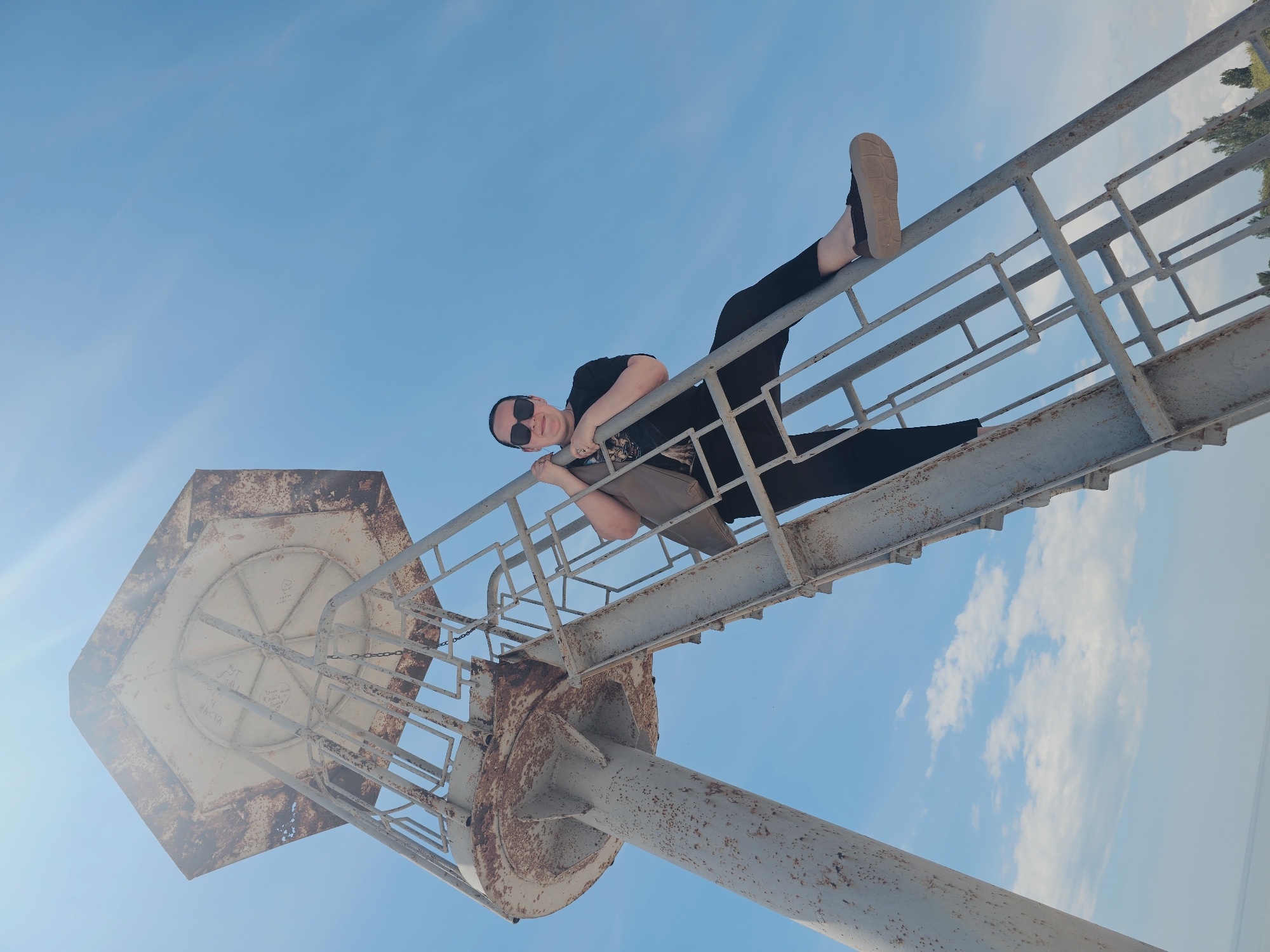 A low-angle, upward-facing photograph captures an individual on a weathered metal navigational structure against a clear blue sky in Skadovsk, Ukraine. The structure, identified as a navigation sign or small lighthouse near the port of Skadovsk, stands predominantly grey-white, exhibiting significant rust and corrosion across its surfaces, particularly at joints and edges. It consists of a sturdy central support column, a circular base platform, and an upper hexagonal or octagonal section, possibly an observation deck or lamp room, with unreadable graffiti visible on its underside. An external, cage-like metal ladder ascends the structure, featuring distinctive rectangular rungs and supports.
A person, appearing to be female, is positioned on this ladder. She wears dark, reflective sunglasses, a black t-shirt with a faint graphic design, black pants, and dark flat slip-on shoes with light soles. A light brown or grey shoulder bag hangs from her left shoulder. She is observed holding onto the ladder's vertical elements with both hands, her left arm hooked over a rung, and her legs bent, with her feet resting on different rungs. Her posture suggests a posed interaction with the structure, and she is looking directly towards the camera with a slight smile.
The environment is dominated by a bright, clear blue sky, with subtle scattered white cumulus clouds visible in the lower right quadrant. Strong sunlight from the upper left creates lens flare and high reflectivity on the metal surfaces. In the distant background, between the structure's supports, a faint horizon line indicates a body of water, consistent with the Karkinit Bay or Black Sea coast on which Skadovsk is situated. The tops of green deciduous trees are visible on the lower right, suggesting nearby land.