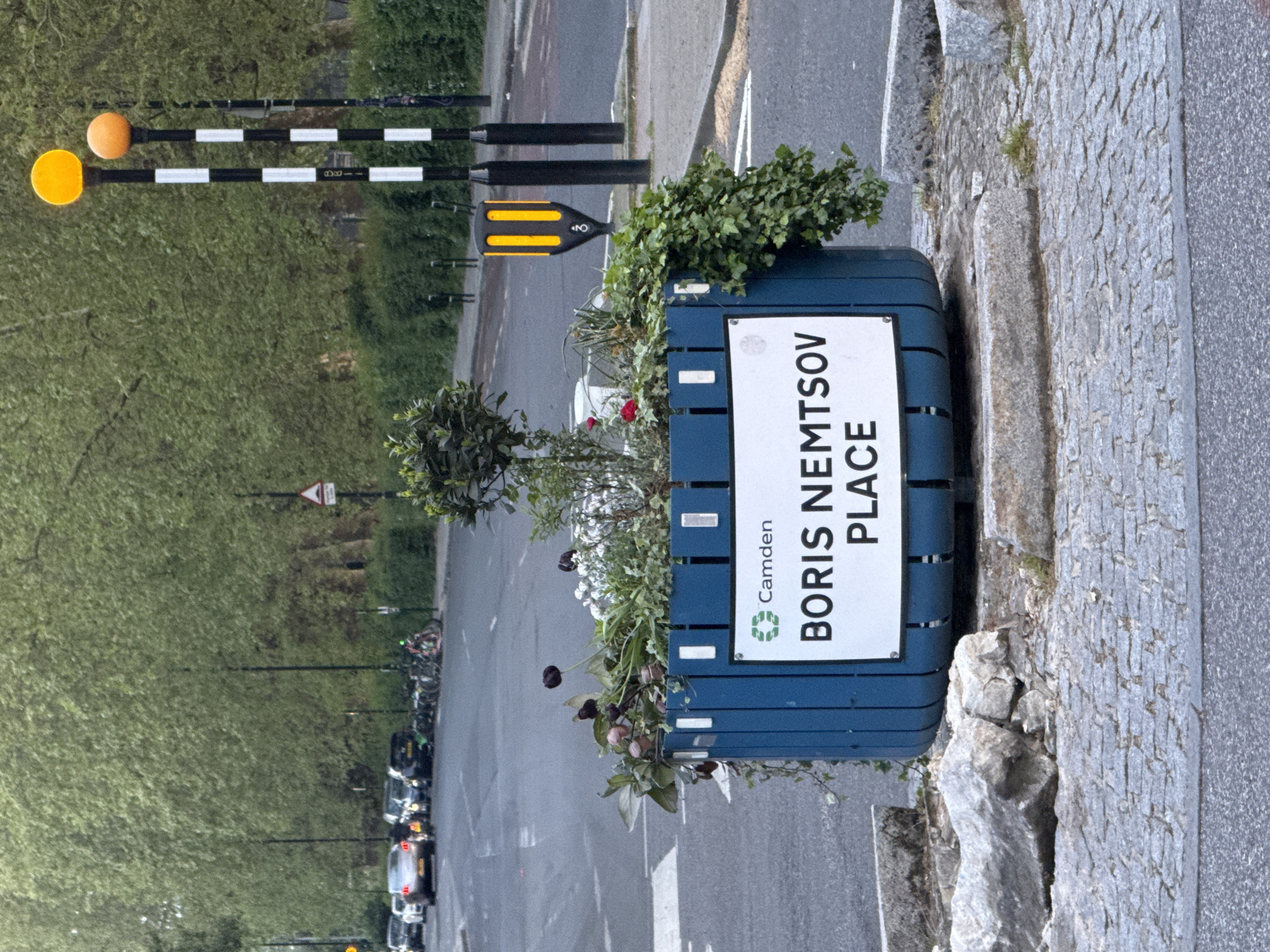 The image shows a blue planter box on a sidewalk, displaying a street sign that reads "Boris Nemtsov Place," indicating that this is a street named after the Russian politician Boris Nemtsov. The sign also includes the Camden logo, suggesting the location is in the London Borough of Camden, United Kingdom. The planter is filled with various plants and flowers. The sidewalk is made of gray stone and borders an asphalt road. Along the road, there are parked cars, street lights, and some street markings. In the background, there is a line of trees, possibly in a park or along the street, and a pedestrian crossing pole with orange globes.