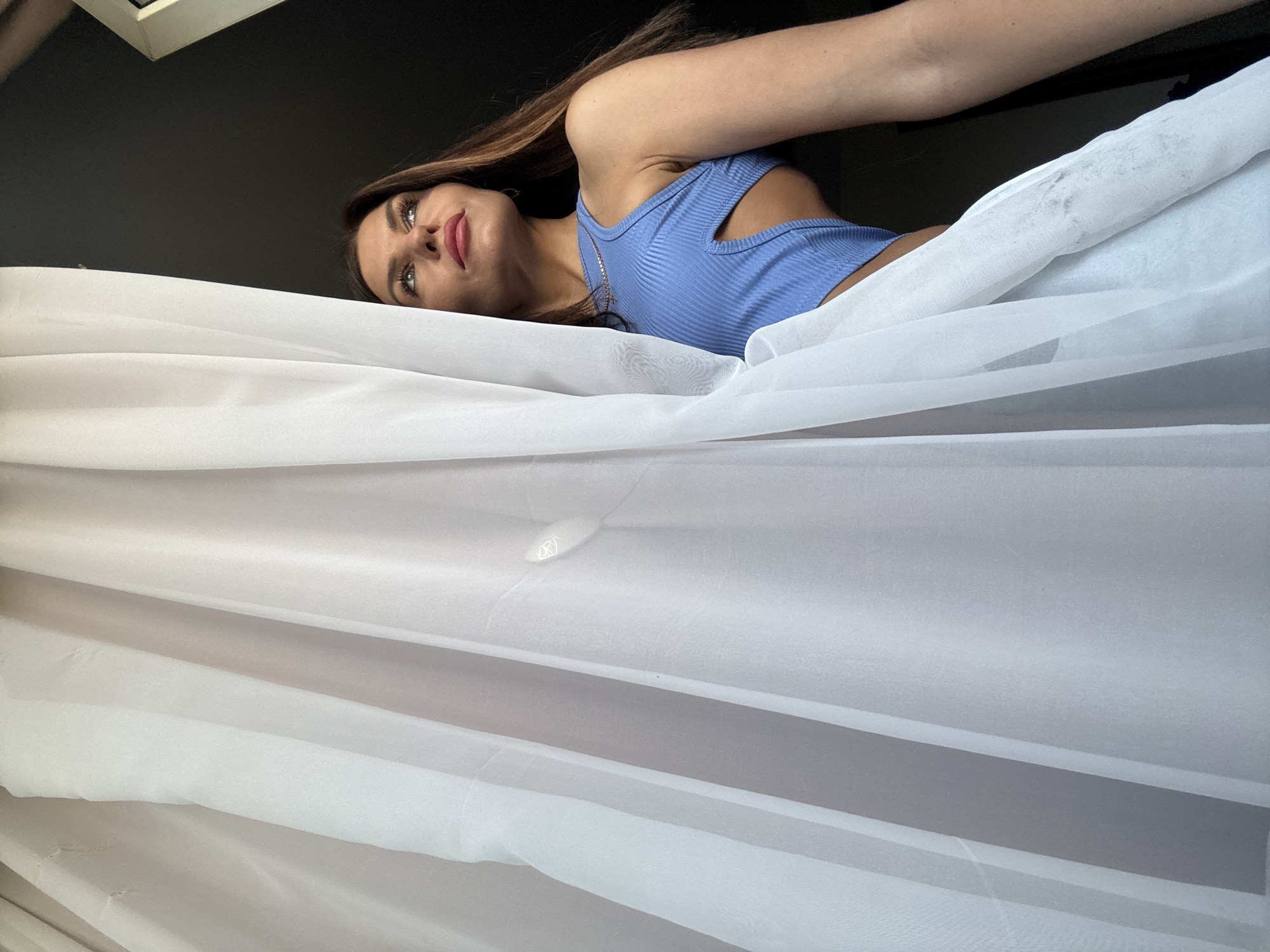A woman with long brown hair is partially obscured by a white sheet. She is wearing a light blue tank top and lying down. The setting is indoors, with a dark ceiling. It is impossible to verify the location in Chaikovsky, Russia from the image.
