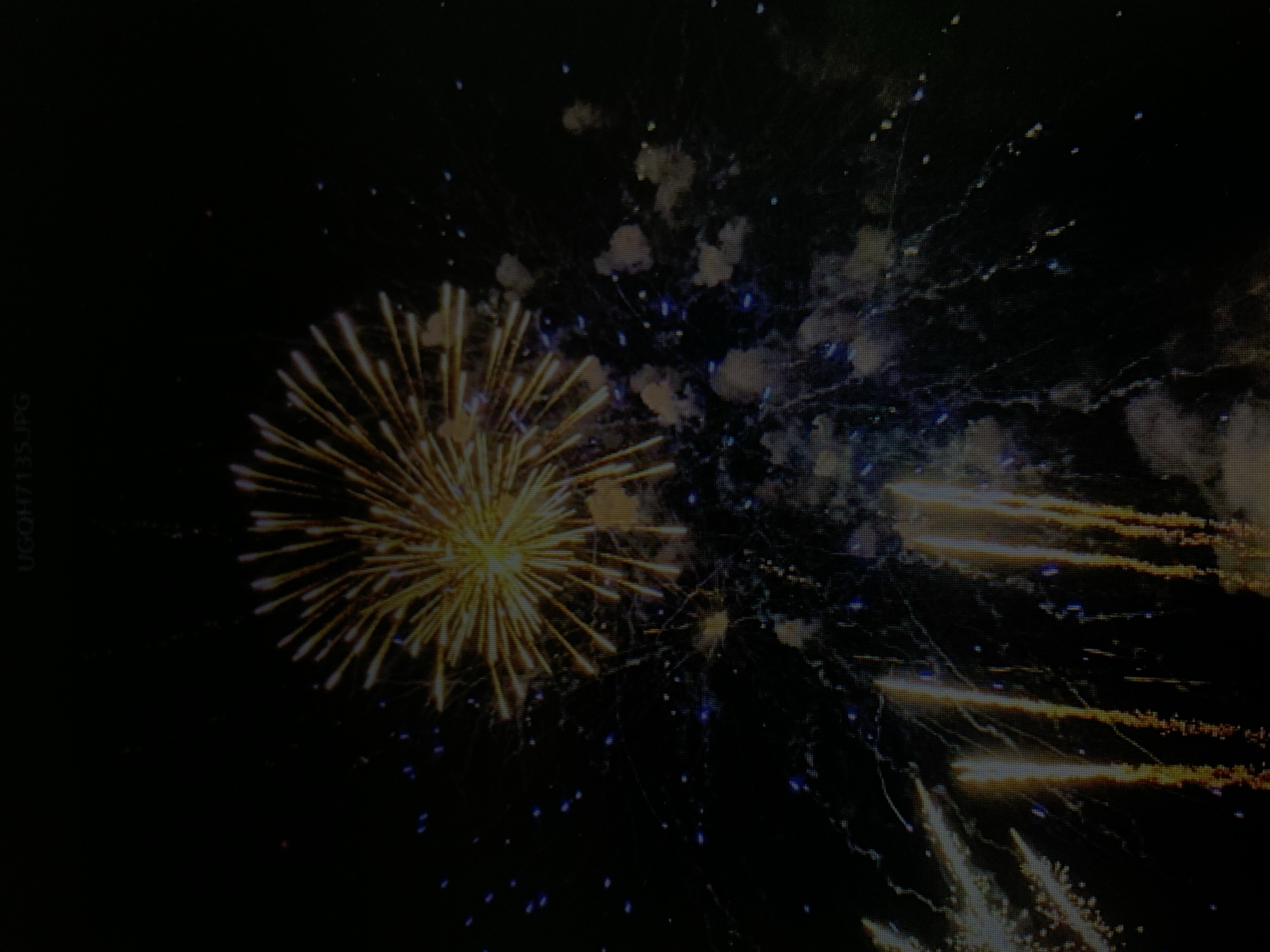 A fireworks display is captured against a dark night sky. A large, bright yellow firework explodes in a radial pattern at the top of the image. Below this, several cascading fireworks are visible, featuring streaks of yellow and orange light. Small blue sparks are scattered throughout the dark background, suggesting the aftermath of other pyrotechnic bursts. The overall scene is one of celebration and festivity. The location is not specified in the image or its context.
