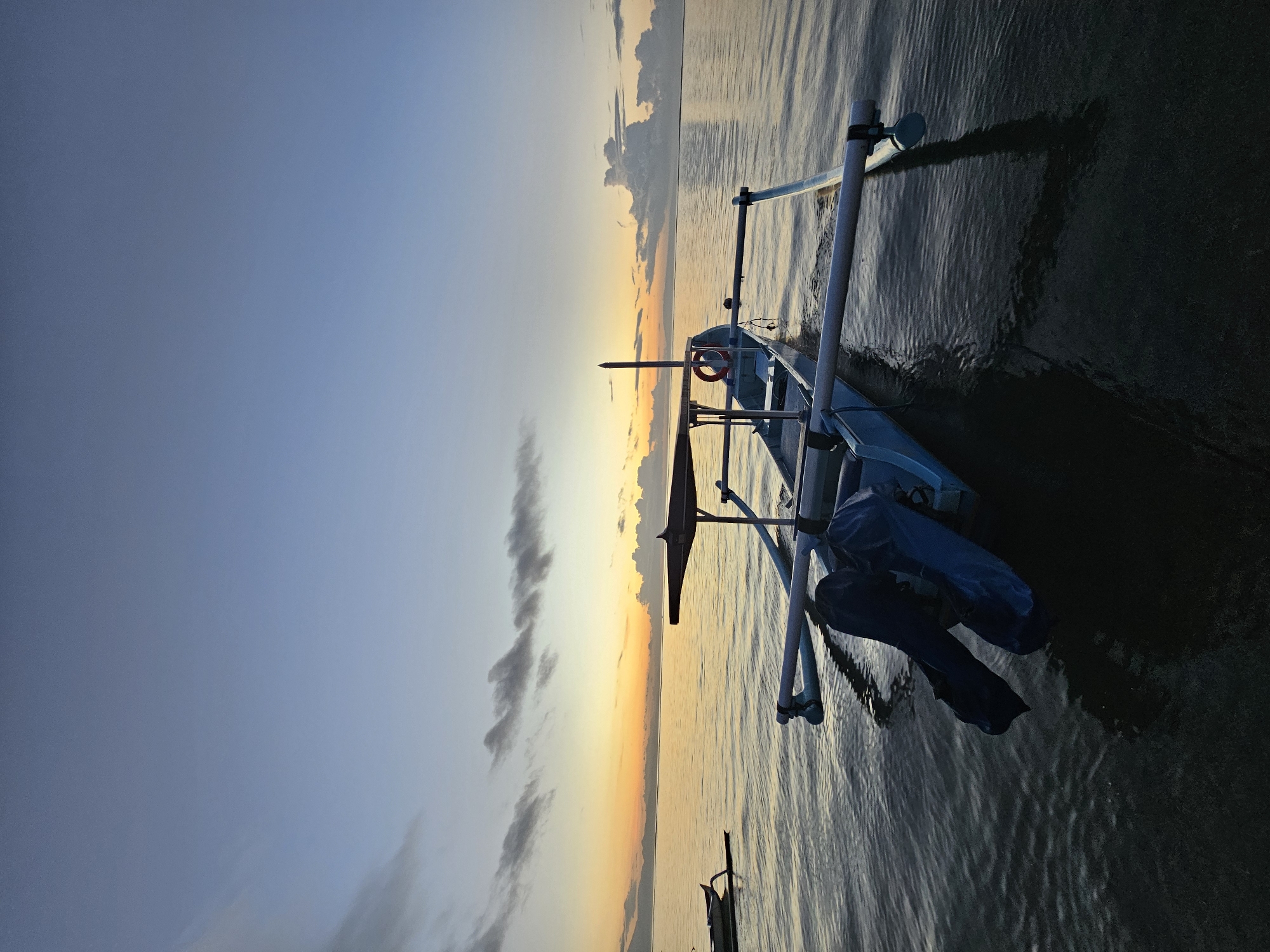 The image shows a traditional outrigger boat at sea at sunset in Denpasar, Indonesia. The boat is light blue with white outriggers. A life preserver is visible on board, and the boat is partially covered with a dark blue tarp. The sky transitions from blue to orange near the horizon. The sea is calm with ripples. There are a few scattered clouds in the sky. The image is rotated 90 degrees counter-clockwise.