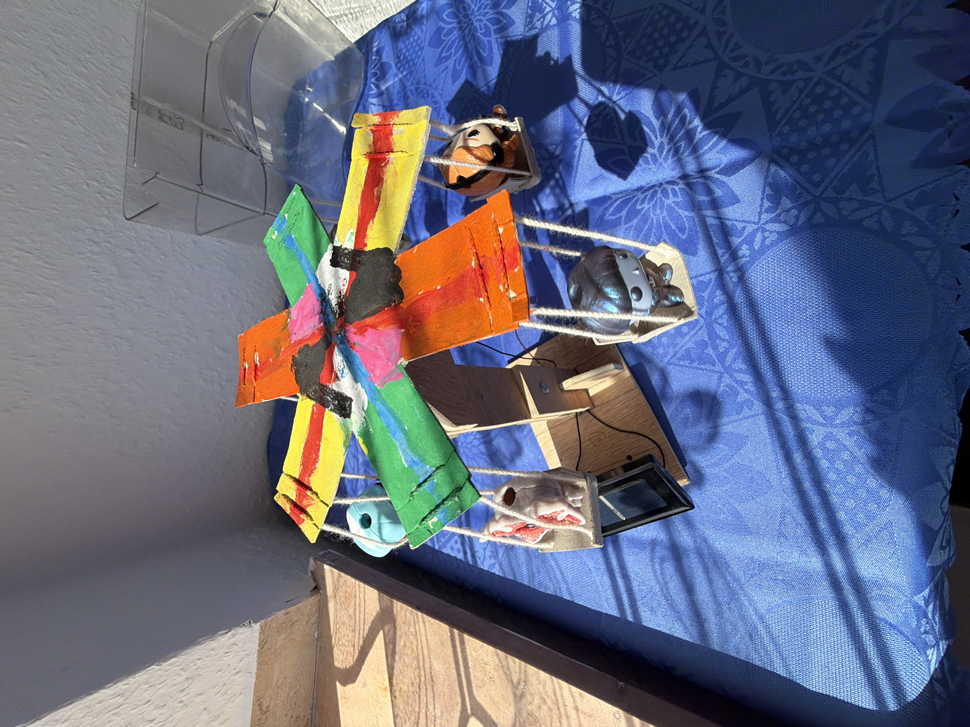 A handmade carousel-like model, painted in vibrant colors including red, yellow, green, and orange, is set up on a blue tablecloth. The carousel has four arms extending from a central wooden structure, each holding a small swing. On the swings are two figurine toys: one appears to be a skeletal figure with red accents, and the other is a dark-haired figure with a metallic sheen. A third swing holds a toy with orange and brown markings, resembling a tiger or cat, and a fourth swing has a turquoise object. The model is illuminated by natural sunlight, casting distinct shadows on the tablecloth. The background consists of a textured gray wall and a portion of a tan tiled surface. A clear plastic container is partially visible behind the carousel. This scene is located in Dorsten, Germany.