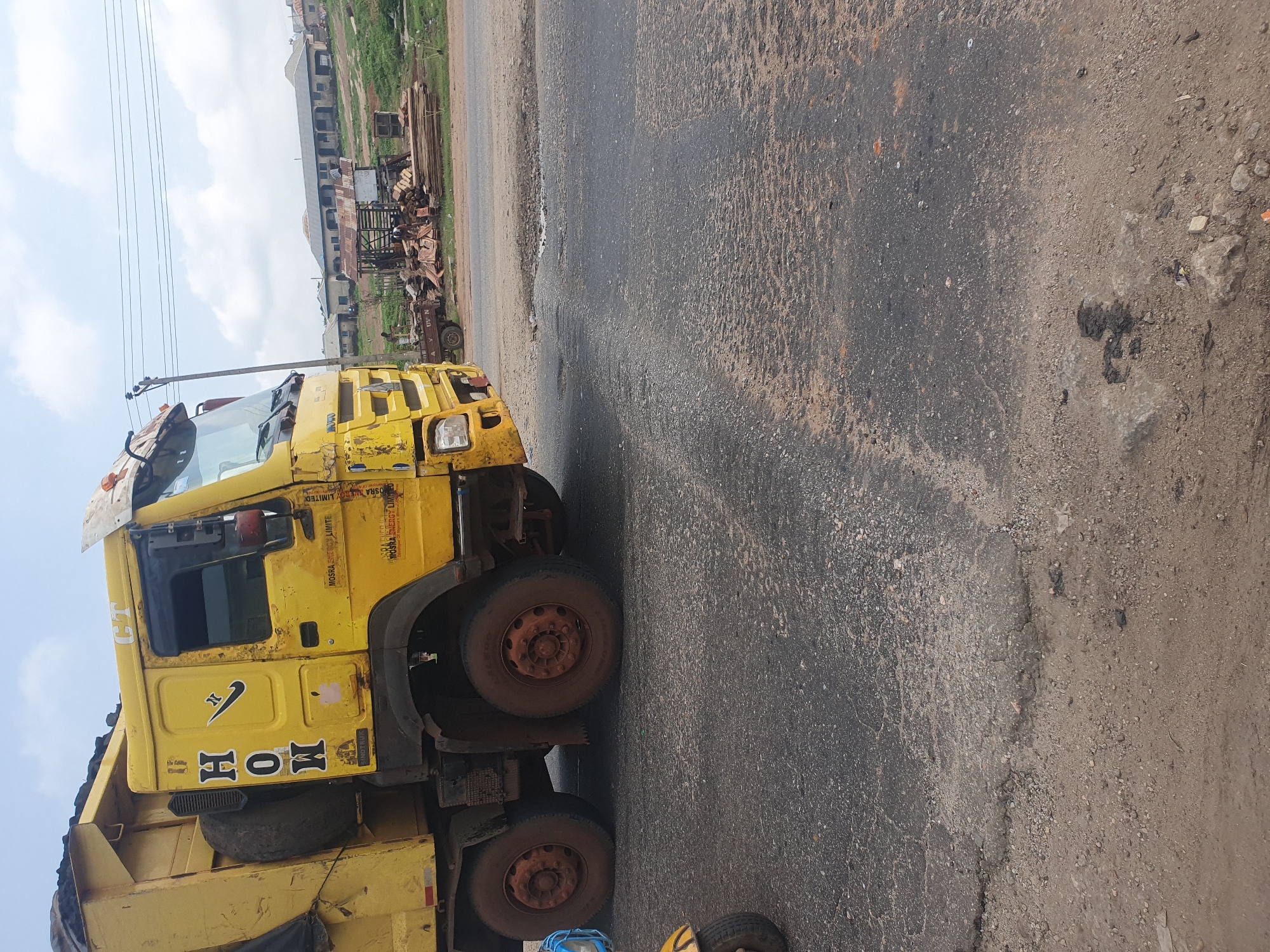An over-the-shoulder, side-view, medium shot captures a bright yellow dump truck parked on a dusty, cracked asphalt road in Lokoja, Nigeria. The truck is filled with dark material in its bed, and the front of the vehicle shows significant wear and tear, with chipped paint and dirt. The words "H O W" are painted vertically in black block letters on the side of the truck's cab, with a black swoosh and a small pink sticker below. Above the driver's side door, "MOSRA ENERGY LIMITED" is printed in yellow text. The truck has large, mud-covered tires.
In the background, several buildings with corrugated metal roofs and what appears to be a construction or lumber yard are visible. A few people are present in the background, with one person standing near a pile of lumber. The sky is overcast with a few patches of blue. The road surface is uneven, with visible potholes and debris. The overall impression is of a working industrial area in Nigeria.