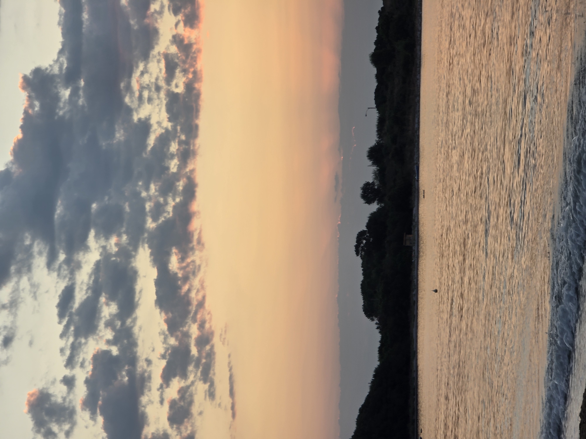 The image depicts a serene seascape at dusk, likely in Chernomorets, Bulgaria. The sky is a gradient of warm orange and pale yellow, transitioning to a soft grey near the horizon, with scattered dark grey clouds edged in pink. A line of dark green trees and vegetation forms a silhouette along the coast. The calm, shimmering water reflects the sky's colors, with gentle waves lapping at the sandy shore in the foreground. A small, light-colored buoy is visible in the water, and a faint structure, possibly a small building or shed, can be seen amongst the trees. A lamppost is also discernible on the distant treeline. The overall atmosphere is peaceful and tranquil, capturing the beauty of the coastal sunset.