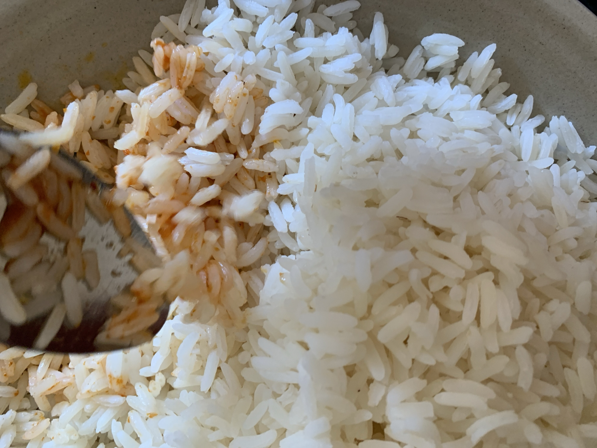 A close-up, overhead view captures a meal of rice served in a light-colored, possibly ceramic or stoneware bowl. The rice is distinctly divided into two sections: the right side and upper-middle portion consist of fluffy, white cooked rice, while the left and lower-middle sections show rice mixed with a reddish-orange sauce or seasoning, suggesting it is either flavored or being mixed with a condiment. A metallic spoon or utensil is partially visible on the left, dipped into the sauced rice, indicating the act of stirring, mixing, or serving the food. The individual grains of rice are clearly discernible. The scene depicts food preparation or serving, likely indoors. There are no people, visible text, or discernible emotions in the image. No specific cues within the image indicate the time of day, weather, or the provided location context of New Stadium, Nigeria.