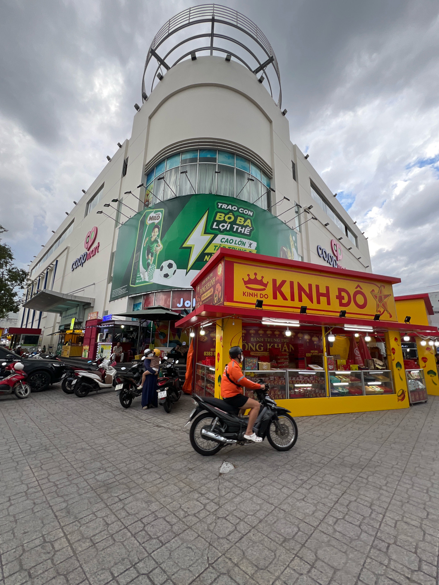 The image shows a street scene in front of a Coopmart supermarket in Nha Trang, Vietnam. The Coopmart building is a large, modern structure with a curved, futuristic design on top. A prominent green billboard for Milo, a chocolate malt drink, is displayed on the side of the building. In front of the Coopmart, a bright yellow and red kiosk sells mooncakes. The kiosk has the brand name "Kinh Do" displayed prominently in large letters.
Several motorcycles are parked on the paved sidewalk in front of the stores. A person wearing an orange jacket and black shorts is riding a motorcycle, moving from left to right across the frame. Another person, a woman in a blue dress, is holding a child in her arms, standing near a parked motorcycle. The sky is overcast with dramatic clouds, suggesting it might be a cloudy day or late afternoon. The street is paved with square stone tiles. There are also other smaller stalls and shopfronts visible, suggesting a busy commercial area. The overall impression is a typical street view in a Vietnamese city, with a mix of modern architecture and bustling local commerce.