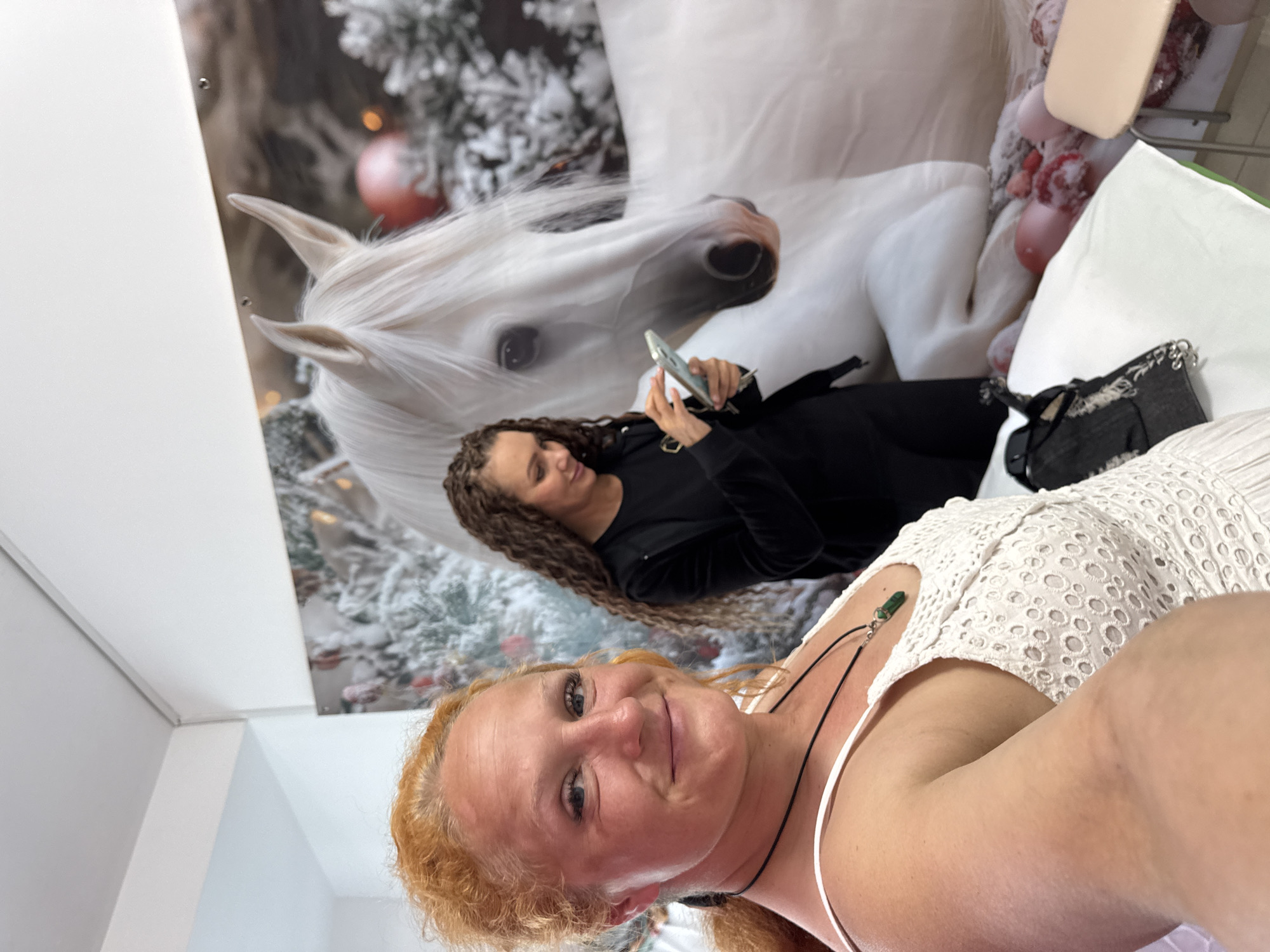 Two women are indoors, posing for a selfie in front of a large mural of a white horse in a snowy, festive setting. The woman in the foreground, with red hair and a white lace dress, is smiling and looking directly at the camera. The woman in the background, with dark curly hair, is wearing a black outfit and looking down at her phone. The mural appears to be on a wall, possibly in a retail space or a decorated room. There are balloons and some soft furnishings visible in the lower right portion of the image. The overall lighting suggests an indoor environment. The image does not provide any visual cues that would allow for its specific location within the city of Chaikovsky, Russia, or any other specific geographical location.