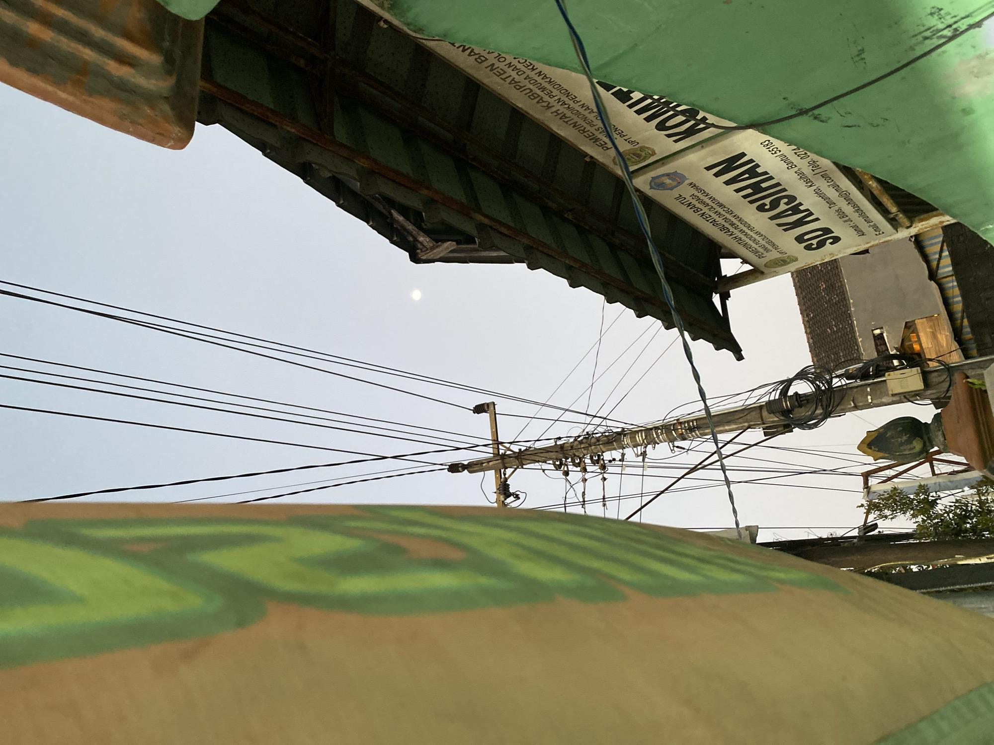 The image depicts a scene in the city of Kasihan, Indonesia, focusing on a street with overhead electrical wires and a sign for "SD KASIHÁN." The sign, displayed on a building with a corrugated metal roof, identifies the institution as "PEMERINTAH KABUPATEN BANTUL DINAS PENDIDIKAN PEMUDA DAN OLAHRAGA UPT PENGELOLAAN PENDIDIKAN KECAMATAN KASIHÁN." The address is listed as "Alamat: Jl. Bibis, Tamantirto, Kasihan, Bantul 55183" and an email address, "email:sdksihan@gmail.com," and a phone number are also visible.
In the foreground, on the left, a large banner or wall with a green abstract design is partially visible. The electrical pole, cluttered with wires and some small black boxes, stands prominently in the center. To the right, the rusted corrugated roof of a building juts out, casting a shadow. The sky above is a pale blue, with a faint moon visible. In the background, houses with tiled roofs can be seen. The overall impression is that of a typical street scene in a semi-urban area in Indonesia, characterized by densely strung electrical cables and local signage. There are no people or animals visible in the frame.