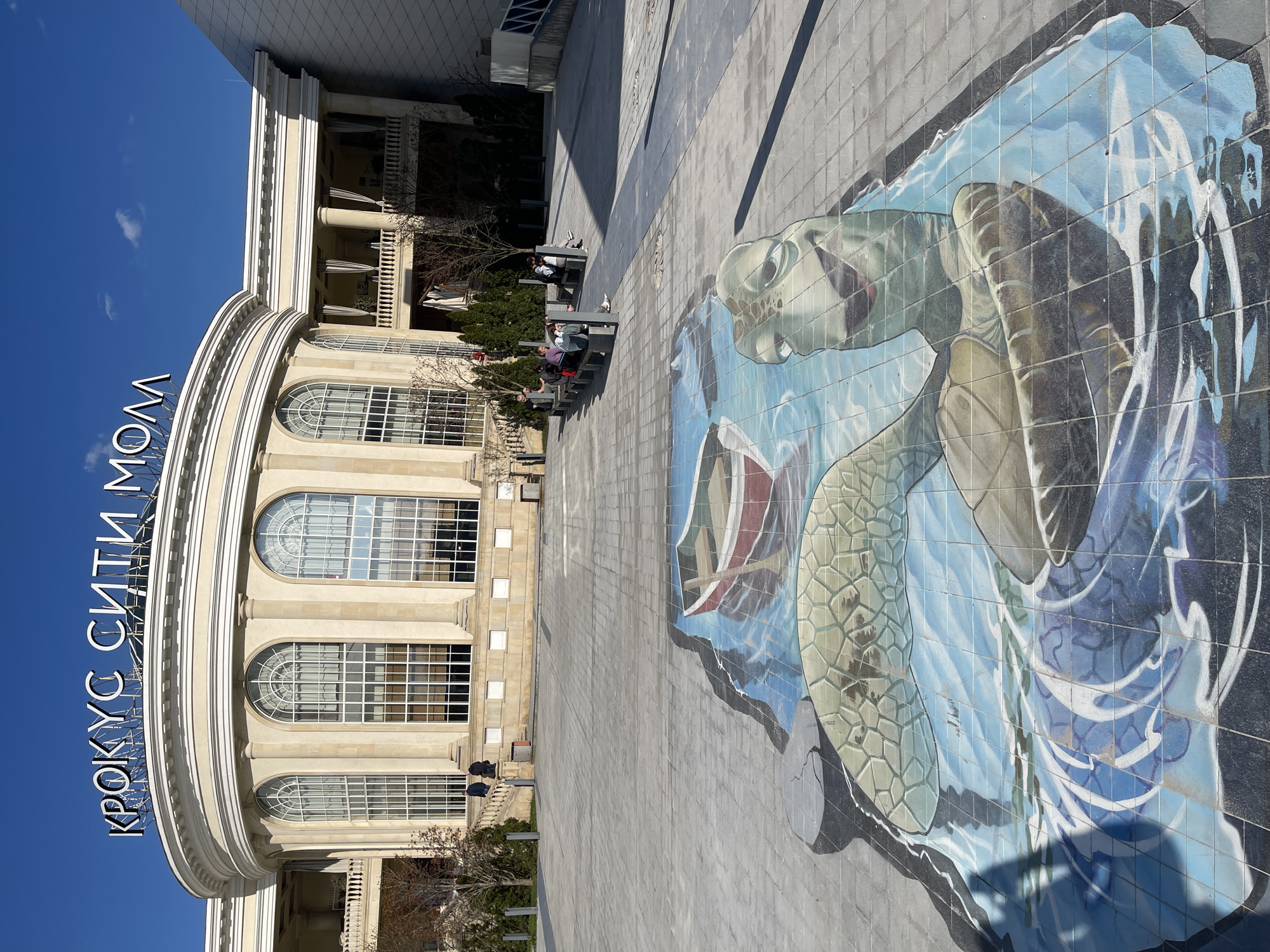 The image depicts a wide shot of a building identified as "Crocus City Mall" located in Khimki, Russia. The architecture is characterized by a curved facade, multiple arched windows, and prominent white columns. Several people are seated on benches in front of the building. A large mosaic of a sea turtle adorns the ground, positioned on the right side of the image. The sky is clear and blue.