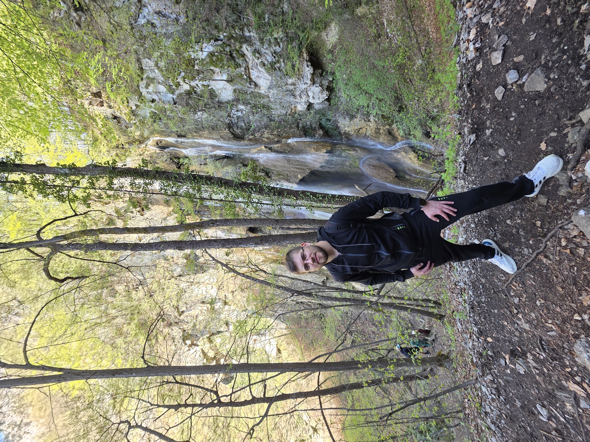 A vertical shot shows a Caucasian man with short brown hair and a beard standing in a wooded area at the Bachkovo Monastery in Bulgaria. He wears glasses, a black tracksuit, and white sneakers. He faces the camera with his hands on his hips. The ground is a mix of soil and rocks. In the background, a waterfall cascades down a rocky cliff face. Trees with budding green leaves surround the area.