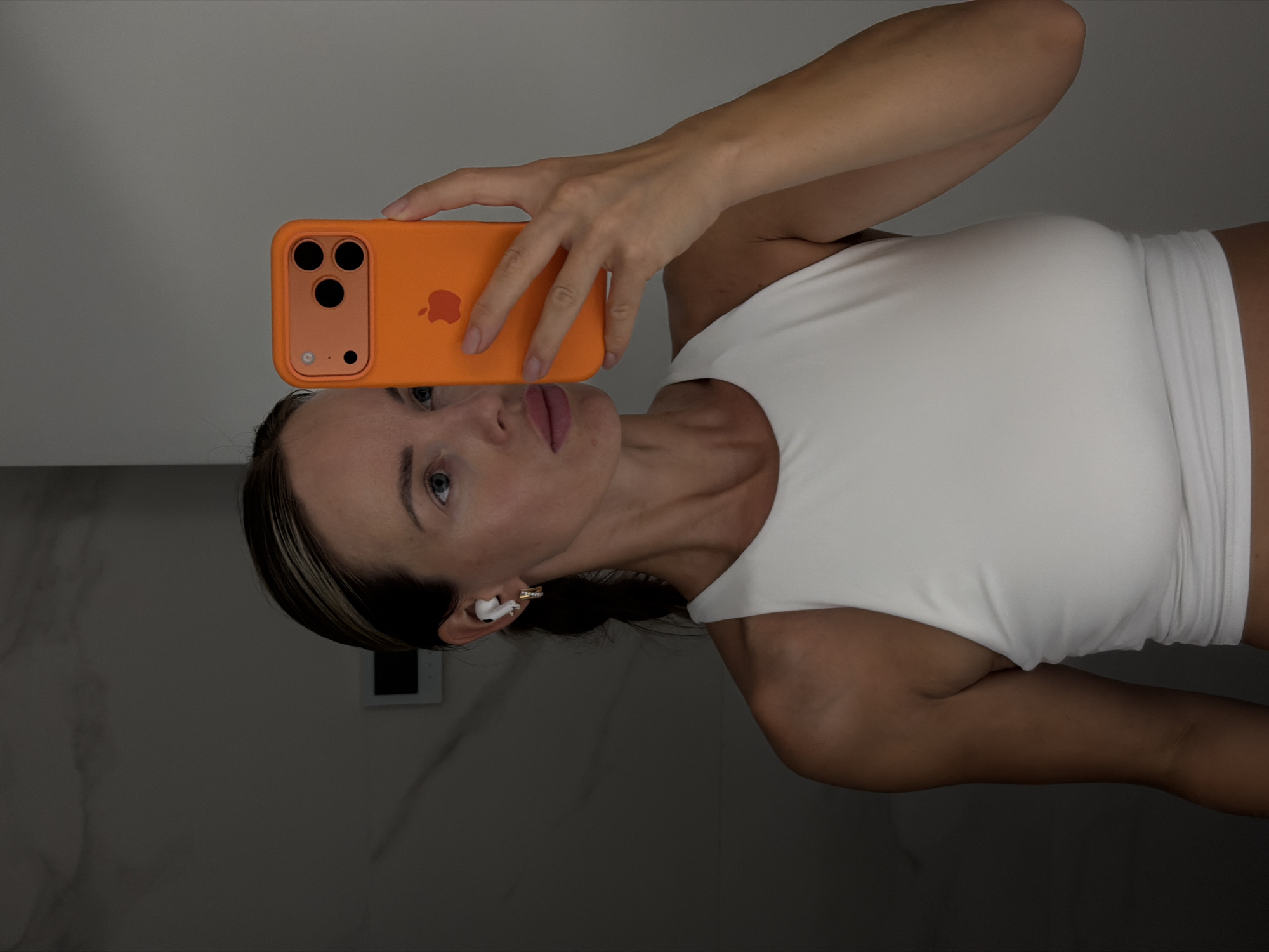 A woman is taking a selfie in a bathroom. She is wearing a white tank top and white earbuds. She is holding an orange iPhone with a case that has an apple logo. The background appears to be a modern bathroom with marble tiles and a light fixture. The image is well-lit, and the woman's face is in focus. The location is not discernible from the image itself.