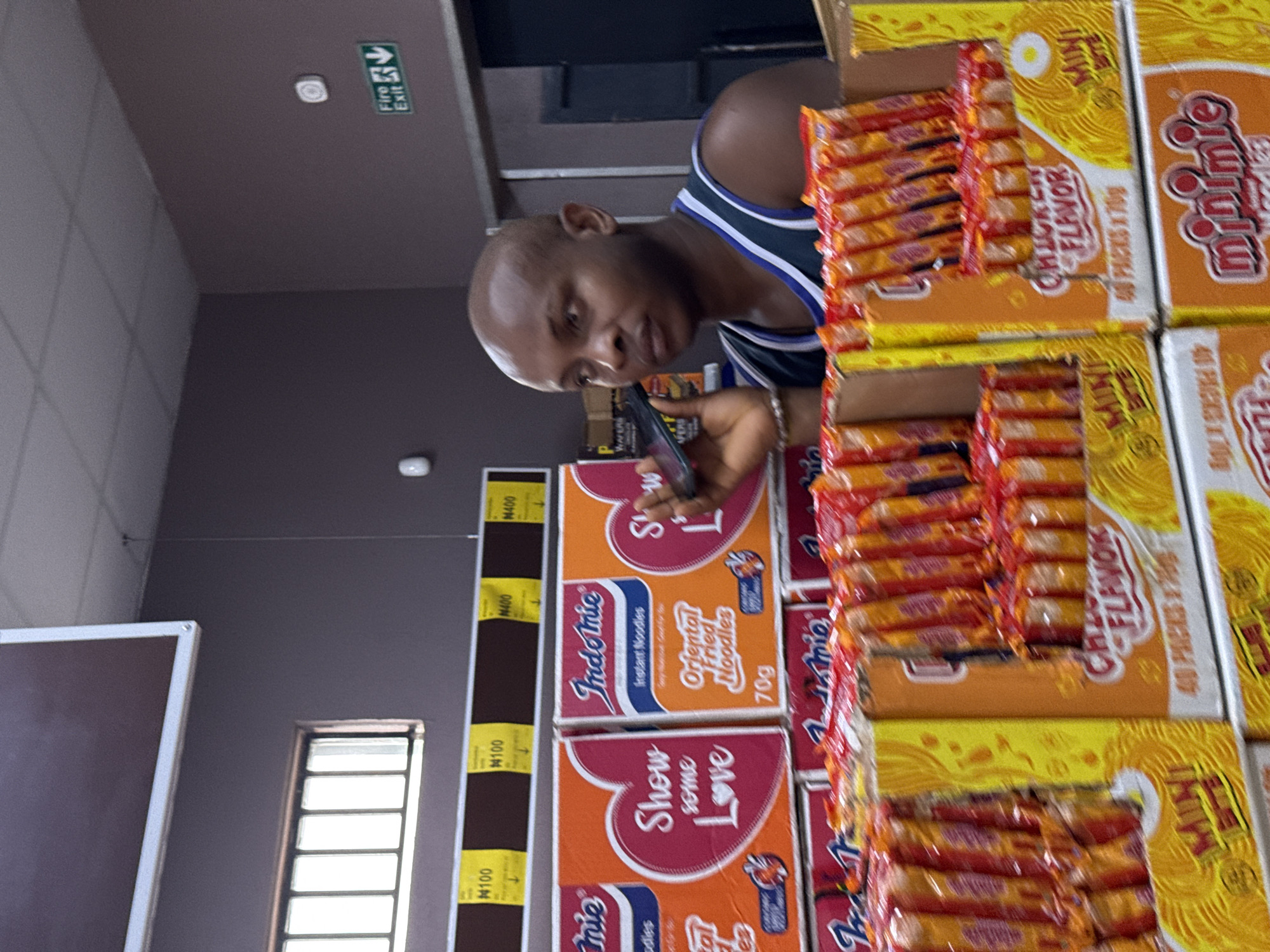 A photograph shows a person using a mobile phone in a store. The individual, a dark-skinned man, is wearing a dark sports jersey with blue and white trim. The man is holding the phone in his right hand, and he appears to be focused on the screen. The store appears to be a shop that sells food products, especially instant noodles. Behind the person, there is a shelf stocked with boxes of Indomie and Minimie brand instant noodles. The boxes have printed text and graphic designs. A yellow price tag is visible to the left of the Indomie boxes with the price N100 and N400. The ceiling of the store is visible with a white fire exit sign. A window is visible at the bottom left.