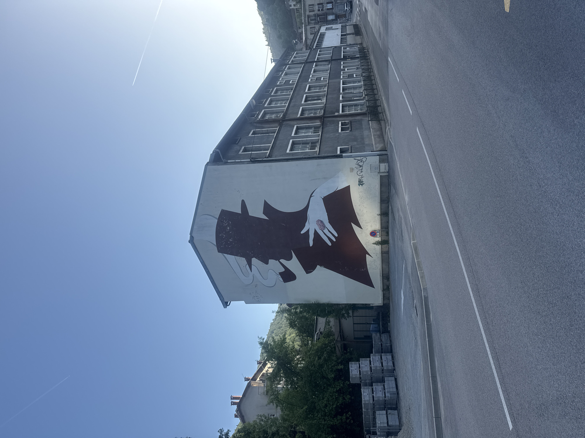 The image depicts a large mural on the side of a building in Saint-Claude, France. The mural is a silhouette of a person wearing a hat and smoking a pipe, with a hand reaching out. The building itself is grey with multiple windows and appears to be an office or apartment building. In front of the building, there is a street with a white line marking the lane. To the left of the mural, there are stacks of grey building materials, possibly bricks or concrete blocks. Trees and other buildings are visible in the background, along with a hilly landscape. The sky is clear and blue, with a faint contrail from an airplane visible. The scene appears to be set during the daytime, with bright sunlight casting shadows.
