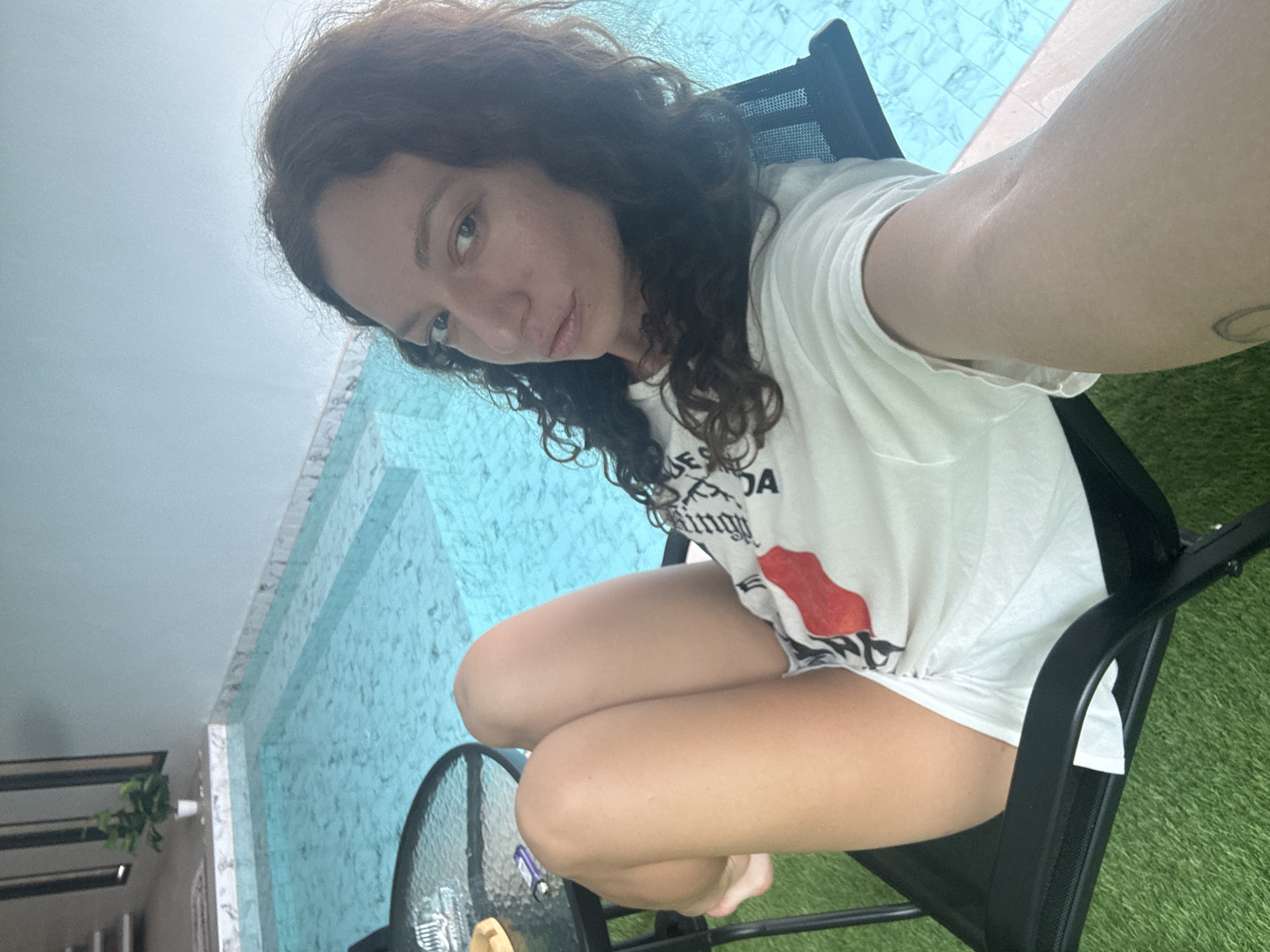 A woman with curly brown hair is seated in a black folding chair outdoors, facing the camera. She is wearing a white t-shirt and white shorts. Her legs are crossed, and her left arm is resting on her lap. To her left, a black glass-top table with a metal frame is visible. The table has some items on it, including what appears to be a lighter. Behind the woman, a swimming pool with blue water and tiled steps is present. The area around the pool is lined with marble. Artificial turf is laid out in front of the chair. The background suggests an outdoor setting, possibly a courtyard or patio area, with a wall and some vertical structures in the distance. The lighting indicates it is daytime. The scene is likely located in a resort or private residence in Ko Samui District, Thailand, given the tropical pool setting.