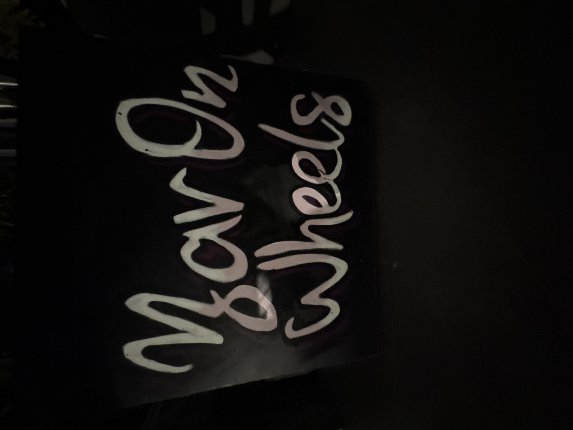 The image shows a black sign with white, stylized lettering that reads "Yar On Wheels". The lettering is slightly reflective and has a pinkish hue in some areas, likely due to the lighting. The sign is positioned in an outdoor, dark environment, with some indistinct background elements that are not clearly identifiable. There are no people or animals visible in the image. The scene is likely in Agios Tychona Community, Cyprus, as indicated.