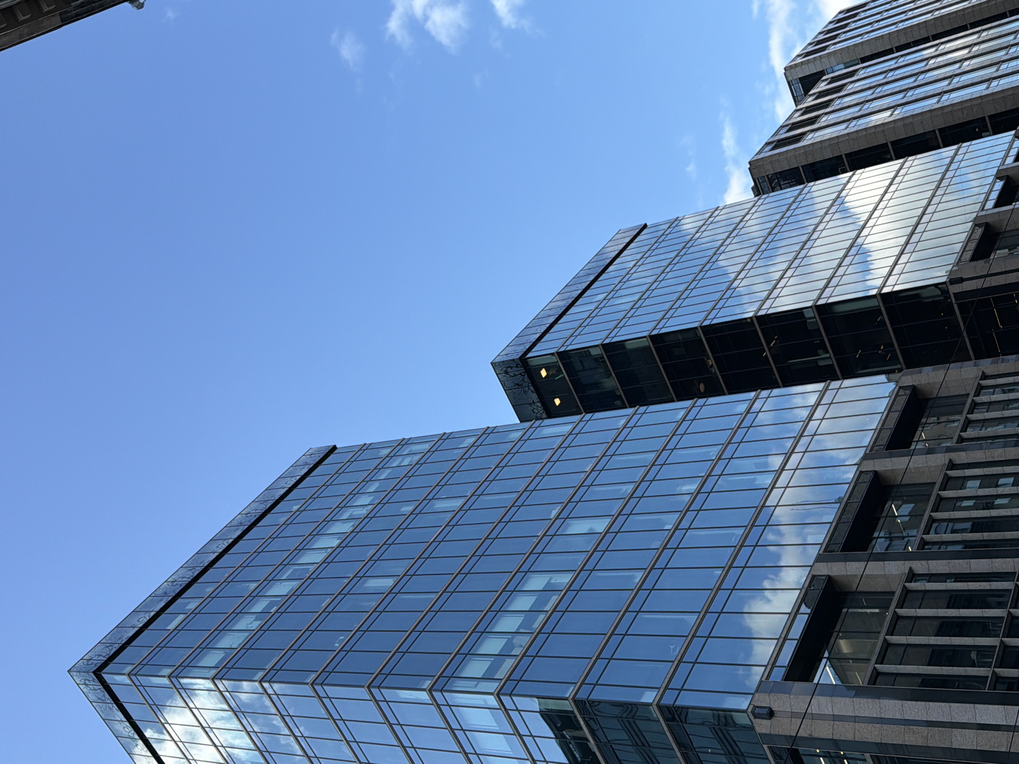 A low-angle view looking up at several modern skyscrapers with glass and dark metal cladding under a bright blue sky with scattered clouds. The buildings feature a grid-like pattern due to the glass panels and support structures. The reflections in the glass panels mirror the blue sky and clouds above. Some of the buildings have more traditional windows in addition to the glass panel sections. The overall scene gives a sense of urban density. The location is unknown.