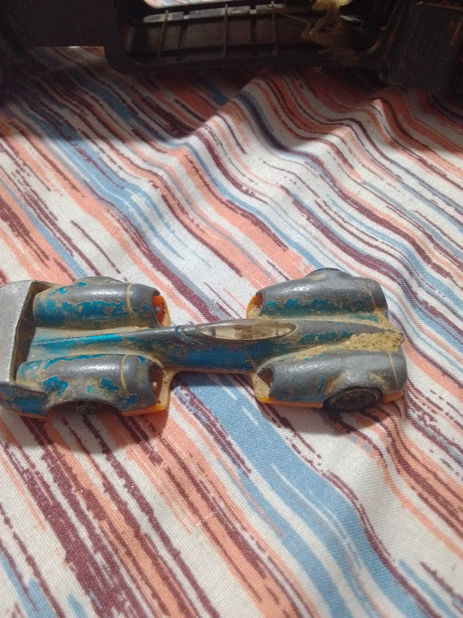 This is a close-up, eye-level shot of a vintage toy car lying on a striped fabric. The car is dark gray with chipped blue paint revealing a metallic base. It has a futuristic, aerodynamic design with two large, rounded engine pods at the rear, each with an orange circular detail. A silver metallic component is visible on the left side of the car, possibly representing a front wing or part of the chassis. The fabric beneath is a textured textile with alternating stripes of white, light blue, salmon pink, and dark reddish-brown. The stripes are diagonal and appear somewhat uneven, suggesting a woven material. The lighting is artificial, casting a soft shadow from the toy car onto the fabric. There are no visible people, text, or specific environmental cues to indicate time of day, weather, or a precise outdoor location, beyond what might be inferred from the setting being indoors on a piece of furniture. The overall impression is one of a discarded or well-loved toy, perhaps in a domestic setting in La Tagua, Venezuela.