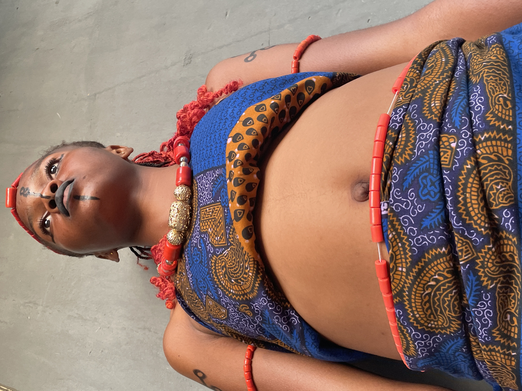 A close-up, high-angle shot captures a person of color, appearing to be a young adult female, lying on a plain, light grey surface, consistent with a concrete floor, in what is likely Badagry, Nigeria. The individual is adorned in vibrant, traditional attire and accessories.
She wears a patterned fabric, possibly a wrapper or shawl, draped around her torso. The fabric features intricate designs in shades of blue, golden-brown, and white/light blue. Her attire is accessorized with numerous red beaded items, including thick, multi-strand necklaces accented with large, ornate gold-colored beads, and matching red beaded bracelets on her arms. Red, coiled or braided hair adornments are also visible around her head and neck area.
Distinctive black markings are applied to her face, notably around her mouth and below her eyes, indicative of cultural body art or makeup. Her gaze is directed upwards, towards the camera, conveying a composed and perhaps solemn expression. The lighting is soft and diffused, suggesting a daytime setting, possibly indoors or in a shaded outdoor area. The person is posing, showcasing her traditional dress and facial adornments. No text is visible in the image.