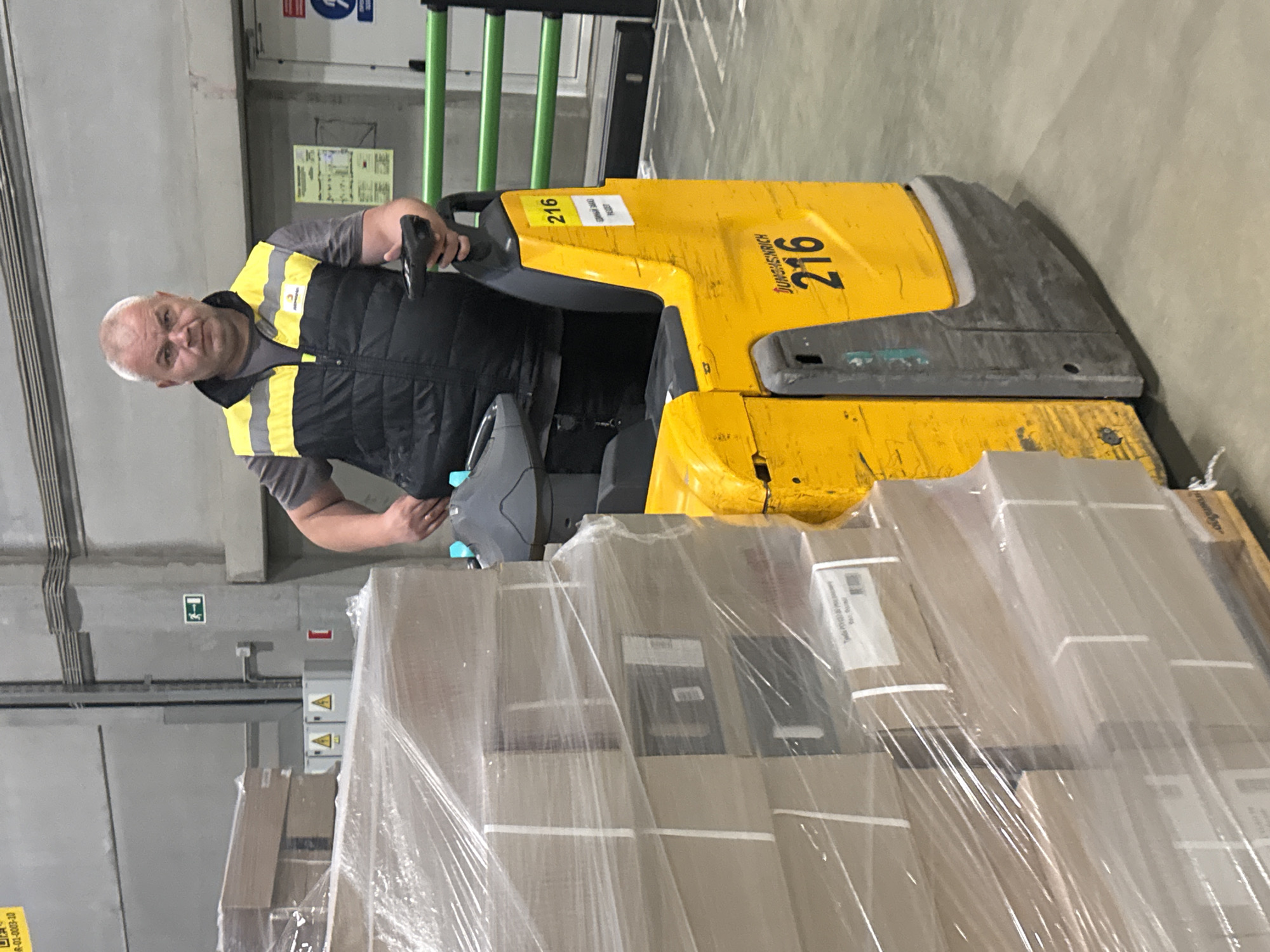 An image, rotated 90 degrees clockwise, captures a man operating an electric pallet jack in an indoor warehouse or industrial facility in Lupanovo, Russia.
The subject is a middle-aged man with short, light grey hair, fair skin, and a neutral expression as he looks towards the camera. He is wearing a grey t-shirt and a black high-visibility vest with bright yellow stripes. His hands are on the controls of the pallet jack.
The man is positioned next to a bright yellow electric pallet jack, manufactured by "Jungheinrich," which is prominently displayed on its side. The number "216" is visible on two separate yellow stickers on the machine, along with partially obscured text on another sticker that appears to read "CEPHA..." and "SAWO..." A tall stack of cardboard boxes, securely wrapped in clear plastic shrink-wrap, rests on a wooden pallet in front of the pallet jack. Some text is visible on the box labels, including "Takis Dydzio Paletas" and "W_R-01-0005-20".
The background features grey concrete walls with some electrical conduits and a green metal railing. A doorway is visible further back. Several safety signs are present on the walls, including a green sign with a white arrow indicating an emergency exit, and a blue circular mandatory action sign with a white symbol above the doorway. The lighting is bright and artificial, typical of a commercial or industrial setting.