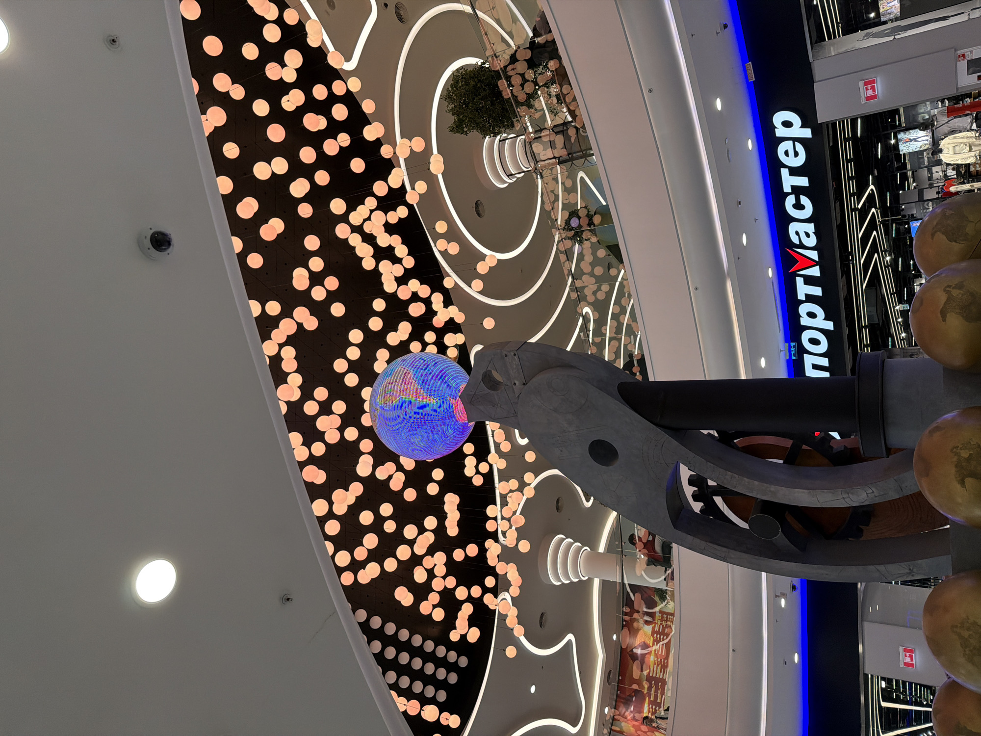 An eye-level, wide-angle shot captures the interior of a multi-level shopping mall. The scene is dominated by a large, gray, abstract sculpture in the foreground, featuring metallic, circular elements and a spherical, illuminated globe at its apex. The sculpture is positioned in front of a large store sign that reads "портМастер" in white, bold lettering with a red "V" symbol integrated into the word "Мастер". Above the sculpture, the ceiling of the mall is decorated with numerous glowing, spherical lights in a warm, orange hue, suspended against a dark background. The mall's architecture features curved balconies, recessed lighting, and a spiral-shaped column in the middle ground. A few people are visible on the balconies and in the background, casually browsing or walking. The lighting is a mix of ambient mall illumination and the decorative lights, creating a vibrant and modern atmosphere. The location is the interior of a shopping mall.