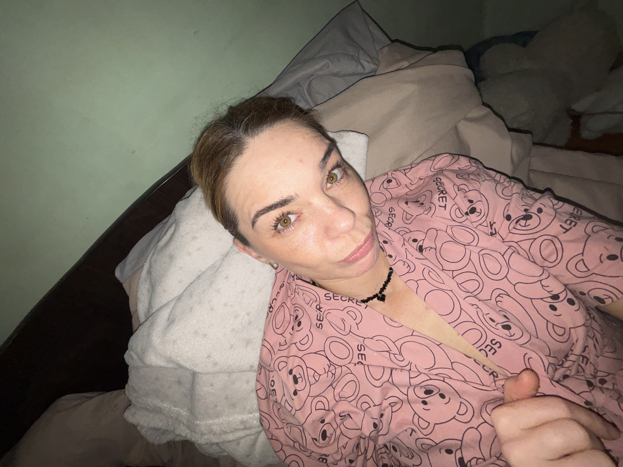 A woman lies on a bed, her head propped up by pillows. She is wearing a pink robe with a teddy bear pattern. Her eyes are open, and she is looking towards the camera. The room is dimly lit, with a green wall behind her. There are no other people or objects visible in the image. The image is a selfie taken by the woman. There is no information in the image to determine the location in the city of Unknown, Russia.