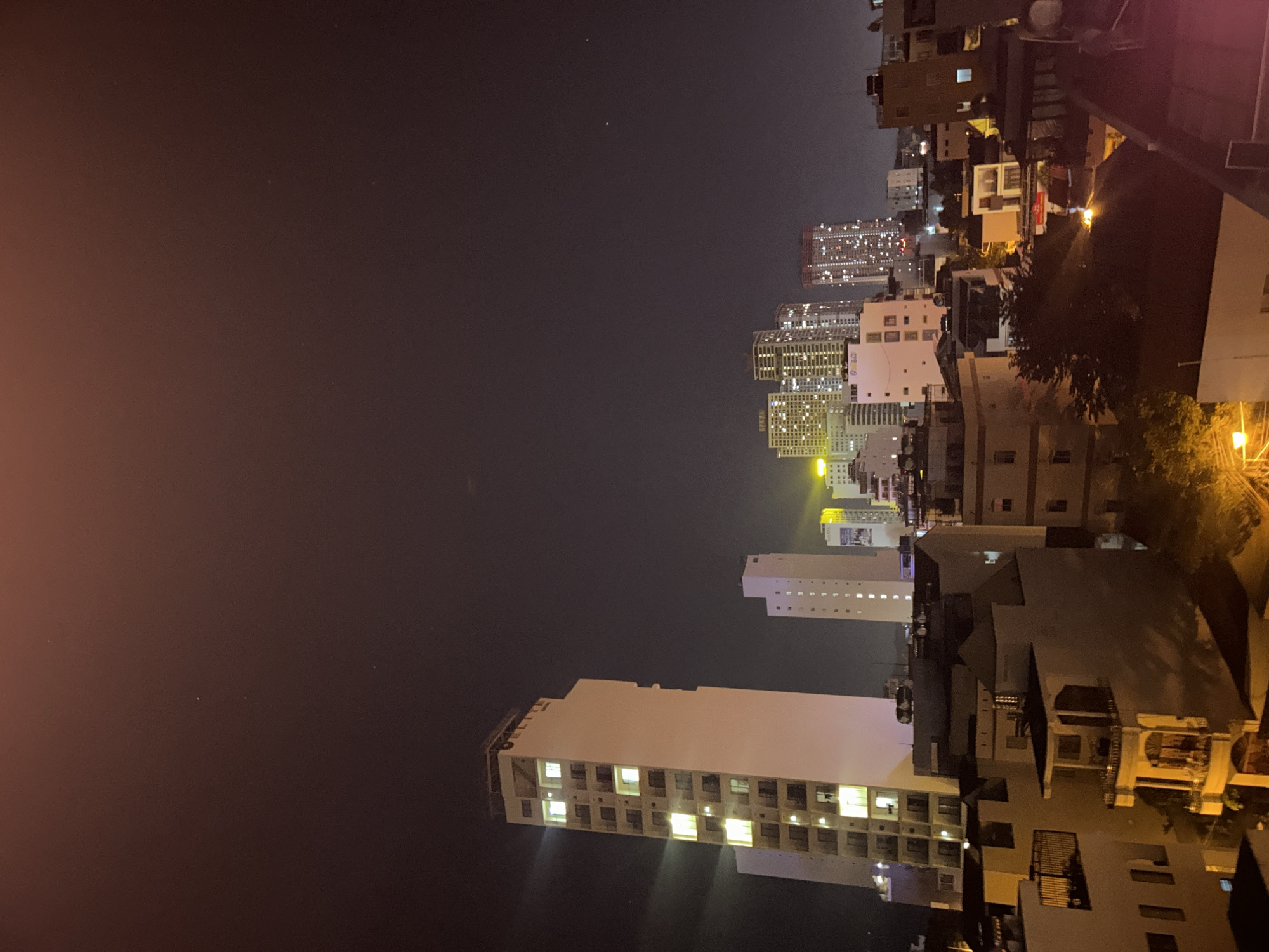 This night-time image presents a bustling cityscape in Bắc Nha Trang, Vietnam, viewed from an elevated vantage point. The scene is dominated by an array of buildings, ranging from multi-story residential or commercial structures to towering high-rises in the distance.
The dark night sky is illuminated by countless artificial lights emanating from the buildings, creating a vibrant glow across the urban landscape. A subtle reddish-orange hue permeates the sky on the left, indicative of city light pollution. In the foreground and mid-ground, several buildings are brightly lit, with numerous windows casting rectangles of light into the darkness. A prominent, lighter-colored building in the mid-ground clearly displays the word "DELIGHT" on its roofline. Further back, clusters of modern high-rise buildings stand tall, their many windows creating a sparkling mosaic. Smaller, older buildings with warm-toned lighting are also visible closer to the viewer.
No specific human activity or people are discernible, but the widespread illumination strongly suggests a lively city environment active after dark. The clear sky indicates good weather conditions, allowing for a broad view of the sprawling city lights. The image conveys the scale and energy of an urban center at night.