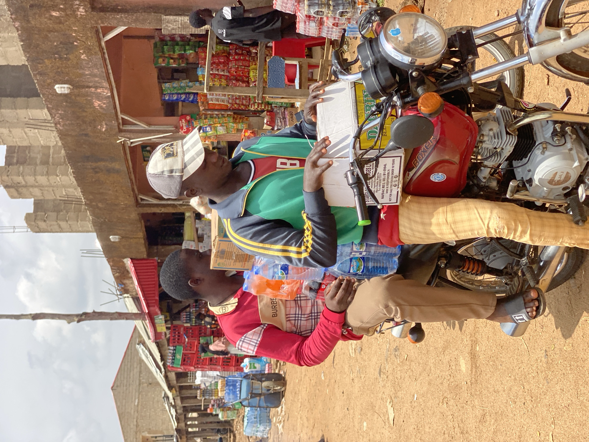 The image depicts a bustling street scene in Kafanchan B, Nigeria, featuring two men near a motorcycle and various market stalls under clear daytime skies. One man, wearing a green and black jacket over a red shirt and a grey and white striped cap, is seated or standing on a red Bajaj Boxer motorcycle. He holds a white box that appears to be a package. The other man, dressed in a red long-sleeved shirt with a plaid pattern (featuring "BURBE" text) and khaki trousers, sits on the motorcycle behind the first man. He wears black "Off-White" branded slides.
The activity suggests a transaction or preparation for delivery, with the men handling goods. The white box on the motorcycle has visible text including "STARCIAGES LI", an industrial address in "Ogus State, Nigeria", a telephone number, "NAFDAC REG. NO.: B1-5994L", and warnings "DRINK RESPONSIBLY: NOT FOR SALE FOR PERSON UNDER 18 YEARS". The motorcycle itself is a red Bajaj Boxer, with "BOXER" and "BAJAJ" visible on its tank and engine respectively.
In the background, several open-air stalls display a variety of products, including snacks, bottled drinks (some "Swan" brand water bottles are visible near the men), and other packaged goods like "JUMBO" and "Cerelac". The ground is dirt, characteristic of unpaved roads or market areas. The overall setting is brightly lit, indicating a sunny day.