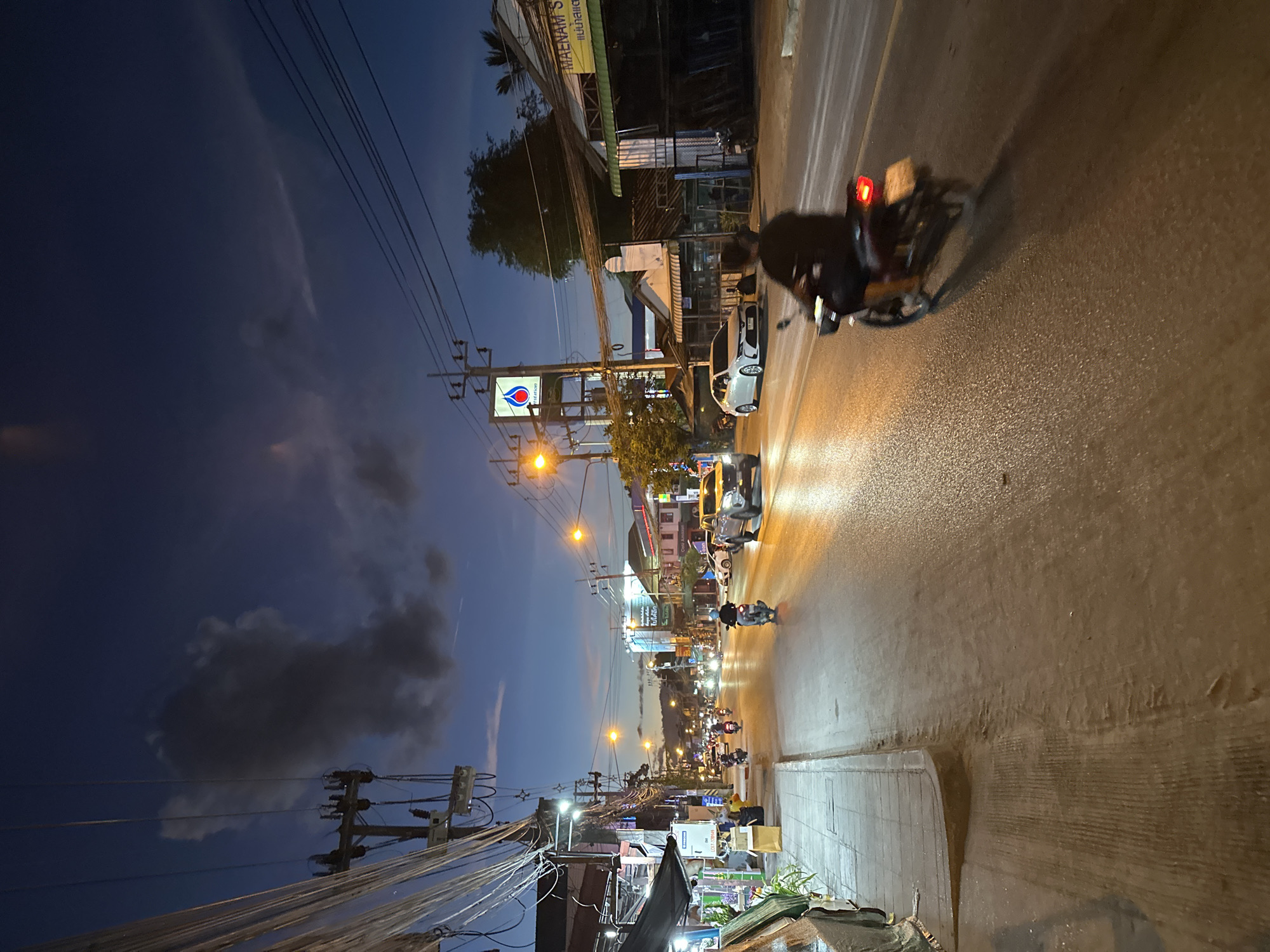 This image captures a street scene in Baan Mae Nam, Thailand, during twilight. The sky displays a gradient of deep blue to lighter shades, with scattered clouds. Overhead power lines crisscross the frame, adding to the urban feel. The street is illuminated by streetlights and the headlights and taillights of vehicles, creating a warm glow on the wet asphalt.
Several motorbikes and a car are visible on the road, suggesting a moderate amount of traffic. One motorbike in the foreground is particularly noticeable with its red taillight illuminated. People are present, though not clearly detailed, as they are either riding vehicles or implied to be within the nearby establishments.
Along the side of the street, buildings with storefronts are visible. One prominent sign displays the text "MAENAM S". Another sign features the iconic red and blue logo of a fuel company, likely a gas station. The overall atmosphere is that of a typical evening in a bustling town, with the transition from day to night in progress. The wet road surface suggests recent rain or ongoing light precipitation, contributing to the reflections of light. The scene conveys a sense of everyday activity and the vibrant life of the location.