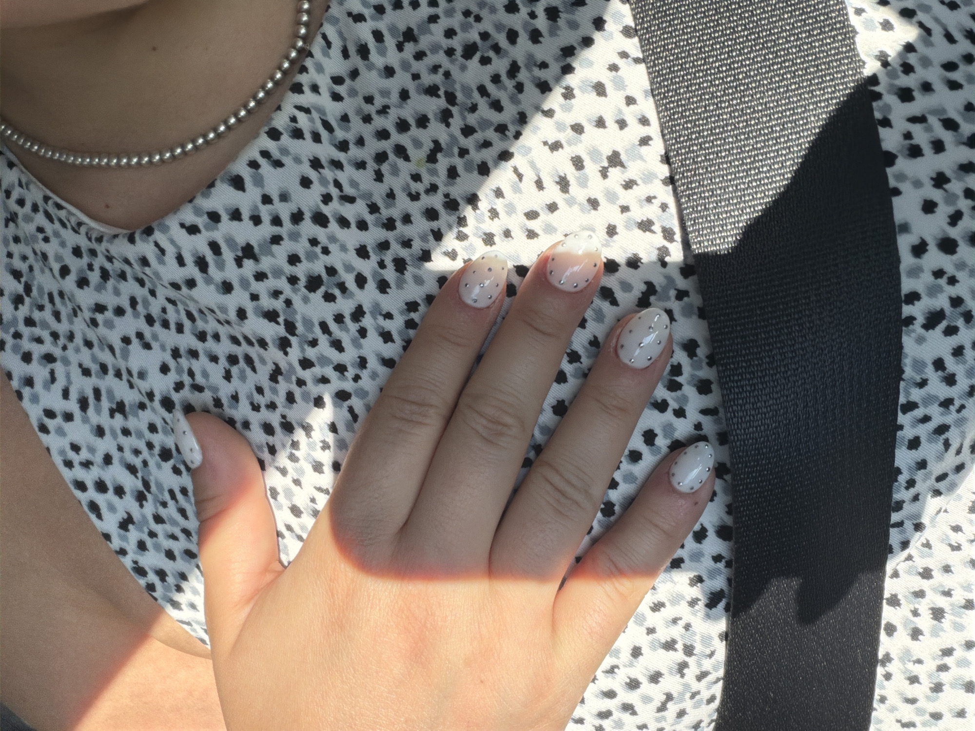 A close-up photograph depicts a person's hand against a patterned fabric backdrop, partially obscured by a seatbelt. The hand is fair-skinned and adorned with nails painted white with small, round, silver embellishments. A beam of sunlight illuminates a portion of the hand. The fabric in the background is printed with a pattern of black and grey spots. A silver beaded necklace is visible near the top left corner of the frame. The seatbelt, made of a textured grey-black material, cuts diagonally across the image from top right to middle left. The setting is likely inside a vehicle, given the presence of the seatbelt. The location is not apparent from this image.
