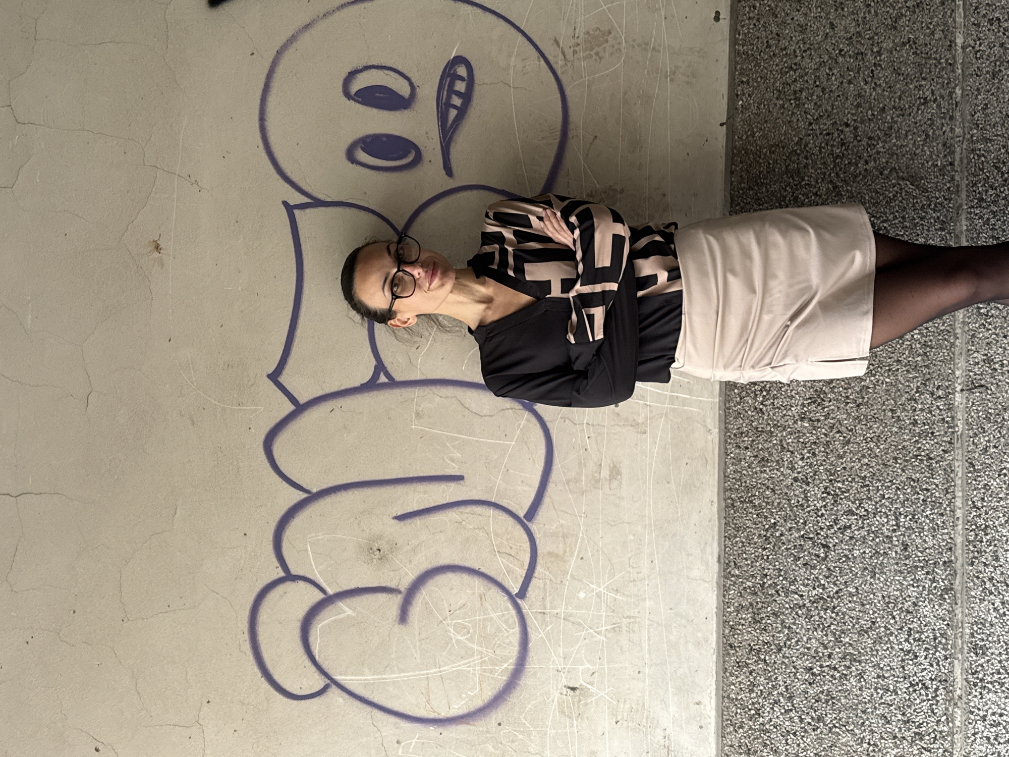 The image features a woman standing outdoors in Pleven, Bulgaria, positioned next to a concrete wall with graffiti. She is captured from the waist up, looking directly at the camera with a neutral to slightly smiling expression. Her arms are crossed, conveying a sense of poise.
The woman wears black-rimmed glasses, a black long-sleeved top with abstract light-brown geometric patterns on the sleeves, a light beige or cream-colored mini skirt, and black sheer tights. Her dark hair is pulled back.
Behind her, on the concrete wall, is a large, stylized graffiti drawing outlined in purple spray paint. The graffiti depicts a cartoonish face with two prominent oval eyes and an open mouth that has a distinct, tongue-like shape on one side. The wall itself shows signs of wear, including cracks and white scuff marks. The ground to her right is a dark gray, textured surface, likely made of small aggregate stones, creating a speckled appearance. The lighting is soft and even, suggesting it is daytime, possibly under an overcast sky or in a shaded area, with no harsh shadows.