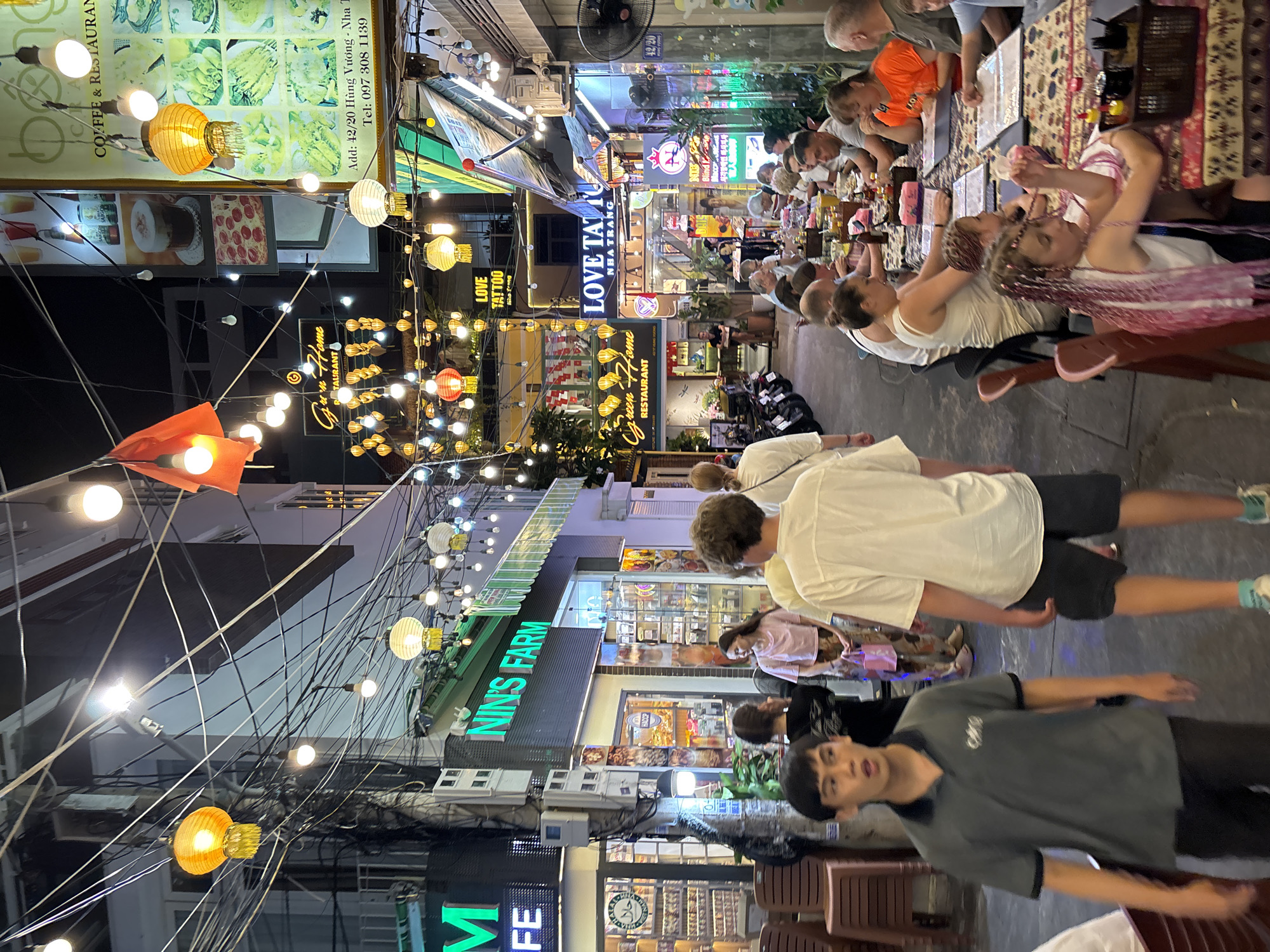 The image depicts a bustling street scene at night in Nha Trang Ward, Vietnam, characterized by a narrow walkway lined with businesses and illuminated by a multitude of decorative lanterns and string lights. The architecture appears to be multi-story, with storefronts and signage clearly visible. One prominent sign reads "NIN'S FARM," while another displays "LOVE TATTOO."
Several people are present. In the foreground, a young man in a grey polo shirt and dark shorts walks towards the camera. Behind him, a person in a white shirt and dark shorts is also walking. Further down the street, a group of people are seated at a long table, seemingly dining outdoors, with food and drinks spread across the table. The lighting is a mix of warm yellow from the lanterns and cooler white from the string lights, creating a vibrant and somewhat crowded atmosphere. The street is narrow, suggesting a pedestrianized area or a lane with limited vehicle traffic, though motorcycles are visible parked along the side. The overall impression is one of a lively commercial and social hub in the city.