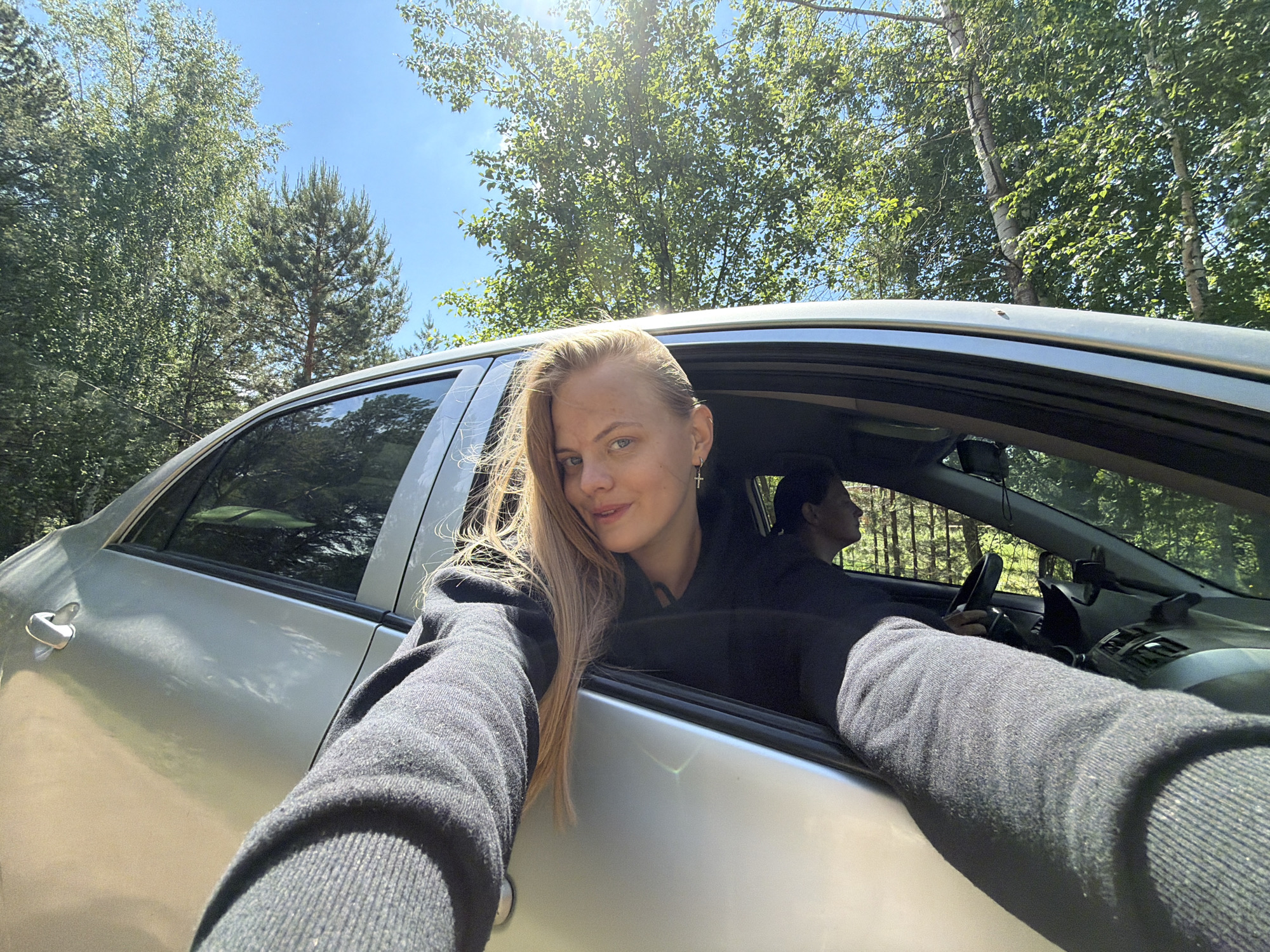 A woman with long blonde hair and a silver cross earring is leaning out of the passenger side window of a silver car. She is wearing a dark gray hooded sweatshirt. The woman is smiling and extending both arms towards the camera, as if taking a selfie. In the driver's seat, a person with short dark hair is visible, holding the steering wheel. The background features green trees and a blue sky with sunlight filtering through the leaves. This image may be taken in the region of Chaikovsky, Russia, given the presence of trees and a typical automobile.