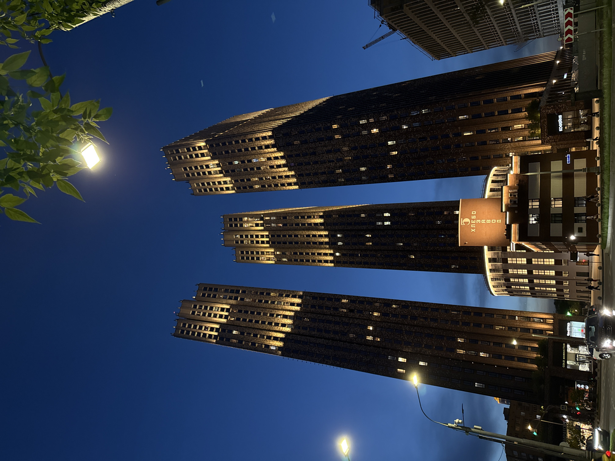 The image captures a view of three skyscrapers in Moscow, Russia, at night. The buildings are tall and modern, with a stepped or tiered design towards the upper floors. Many windows are illuminated, creating a grid-like pattern across the facades. Streetlights are visible, and there are signs in Russian. A construction crane can be seen near one of the buildings. There is foliage in the foreground. The sky is a dark blue.