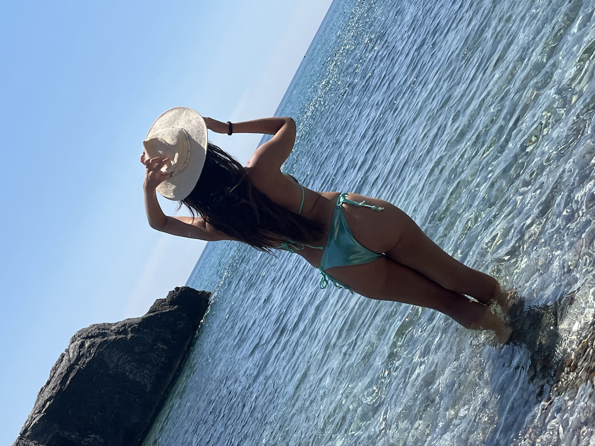 A woman stands in shallow, clear, blue water near a rocky shore. She is viewed from behind, wearing a turquoise bikini and holding a straw hat above her head. The water is calm with small ripples. A large, dark rock formation is visible to the left of the frame, and the sky is clear blue. The scene appears to be a beach in Zagrađe, Montenegro.