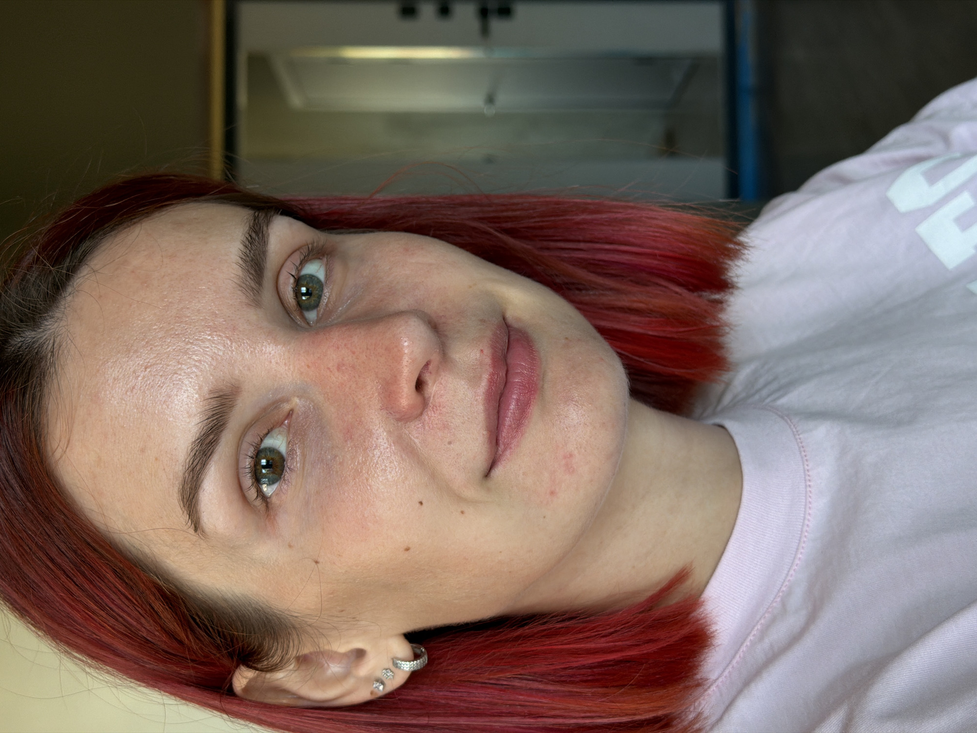 A close-up, slightly angled, indoor photograph features a light-skinned woman with red hair. Her eyes are hazel, and she has light eyebrows. She is wearing a light pink t-shirt. She has several small earrings in her left ear. The background is blurry, with what appears to be a cabinet or shelf unit. The location and city are not discernible from the visual data.