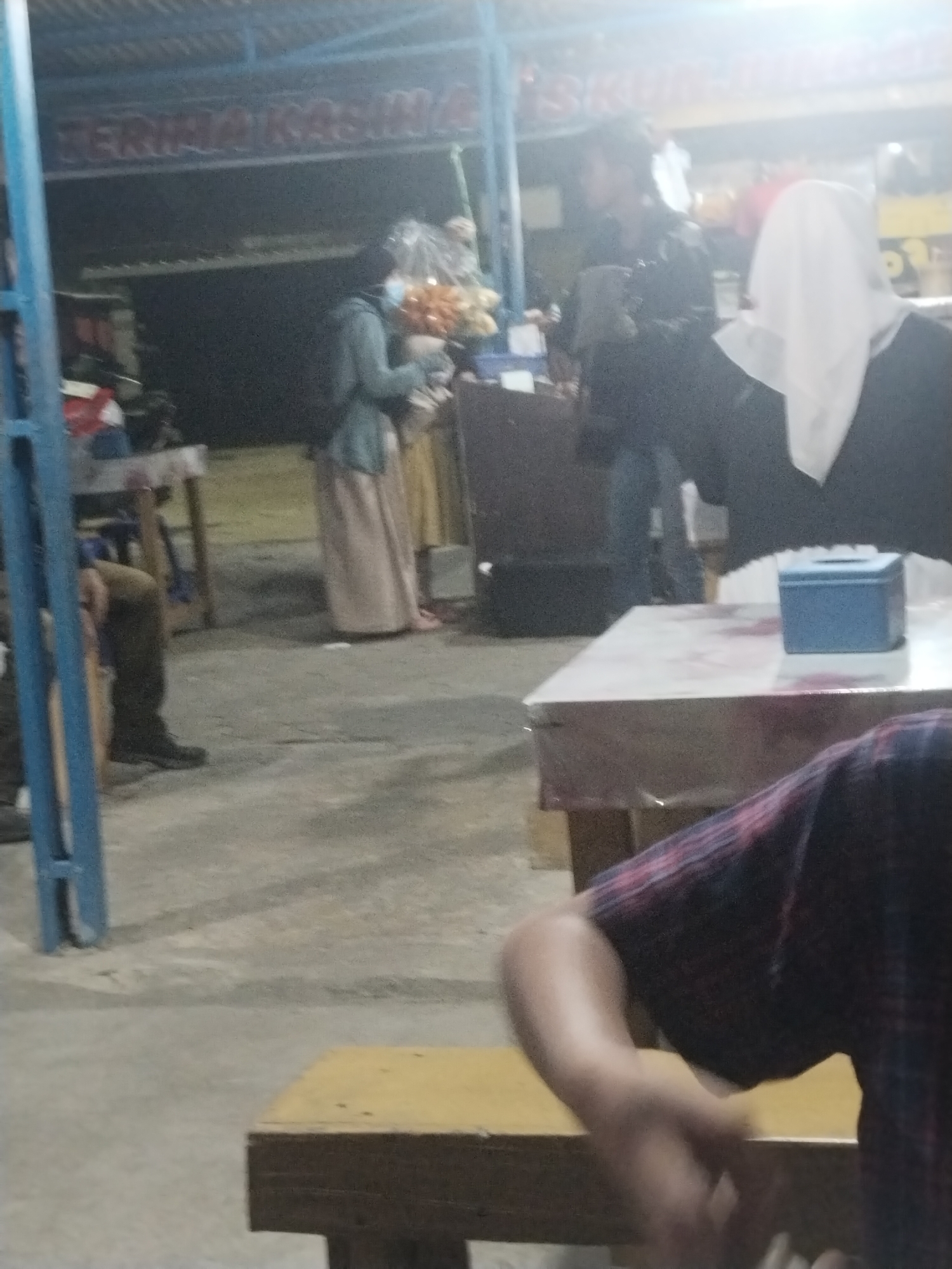 The image depicts an outdoor scene at a roadside establishment in Muara Bulian, Indonesia, likely a food stall or small eatery. In the background, a sign reads "TERIMA KASIH" which translates to "Thank You" in Indonesian. A woman in a light blue hijab and a long, pale pink skirt stands to the left, interacting with someone whose face is obscured by a white hijab. To their right, a person in a black jacket and blue jeans is visible. In the foreground, a person wearing a plaid shirt is seated, their arm and hand blurred as they move. A light-colored table with a pinkish patterned covering is in the middle ground, with a small blue container on it. Another wooden bench is visible in the lower right portion of the image. The ground appears to be concrete or asphalt. The overall lighting suggests it is either dusk or nighttime, with artificial lights illuminating the area.