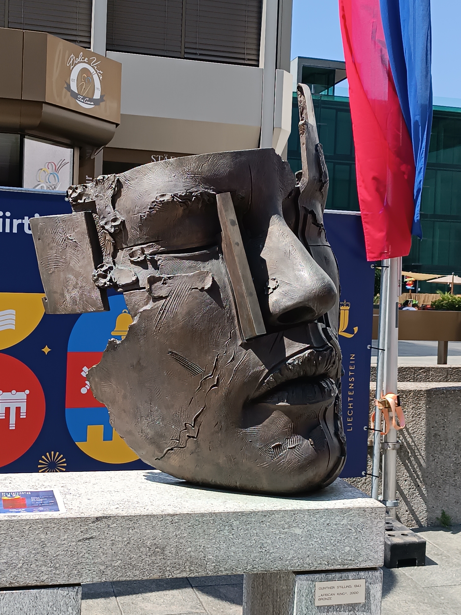 An outdoor, eye-level, medium shot captures a large, abstract bronze sculpture of a face in the city of Vaduz, Liechtenstein. The sculpture is positioned on a granite pedestal. To the right of the sculpture, a metal pole supports two national flags, one red and one blue, fluttering in the breeze. Behind the sculpture, a blue banner displays the coat of arms of Liechtenstein, the word "LIECHTENSTEIN" in white capital letters, and other graphic elements. To the left of the sculpture, a building with a brown facade features a sign that reads "Dolce Vista" above a logo. The background reveals more of the building, a green glass-fronted structure, and awnings of an outdoor seating area. The ground appears to be paved. A small plaque at the base of the sculpture reads "GUNTHER STILLING 1943 'AFRICAN KING' 2000 BRONZE."