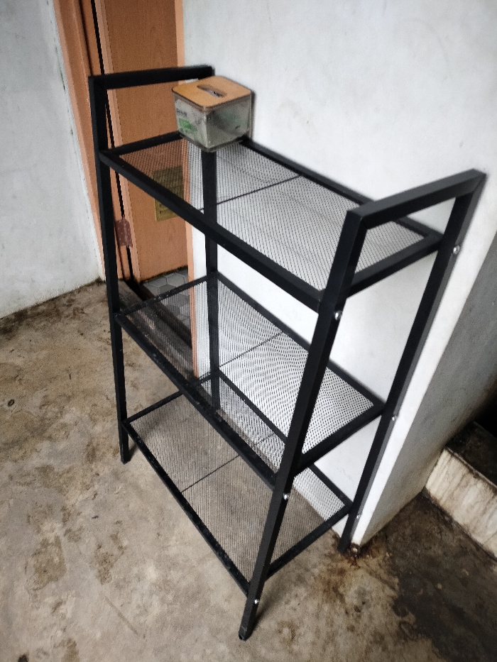 The image displays a four-tiered shelving unit made of black metal with mesh shelves, positioned against a light-colored wall. The unit is angled slightly, with its left side closer to a partially visible brown or orange door frame.
On the top shelf of the unit, there is a small, translucent or clear box with a light-colored, possibly wooden, lid. The lid features a narrow slot, suggesting it might be a donation box, tissue holder, or a small money box.
The immediate surroundings feature a rough, possibly concrete, floor that appears dirty and stained in various places, indicating wear or exposure to the elements. The wall behind the shelving unit is plain and light-colored. To the right of the unit, another wall or structure is visible, also light-colored but with noticeable discoloration and stains at its base, consistent with the condition of the floor.
No specific activity is taking place; the image is a static shot of the furniture in its setting. The lighting suggests daytime, but the exact time or weather conditions are not discernible. The overall scene conveys a functional, utilitarian space, possibly a storage area, garage, or a covered outdoor entrance in Dramaga, Indonesia, given the robust, practical nature of the shelf and the worn appearance of the floor and walls. There is no legible text visible on any of the objects.