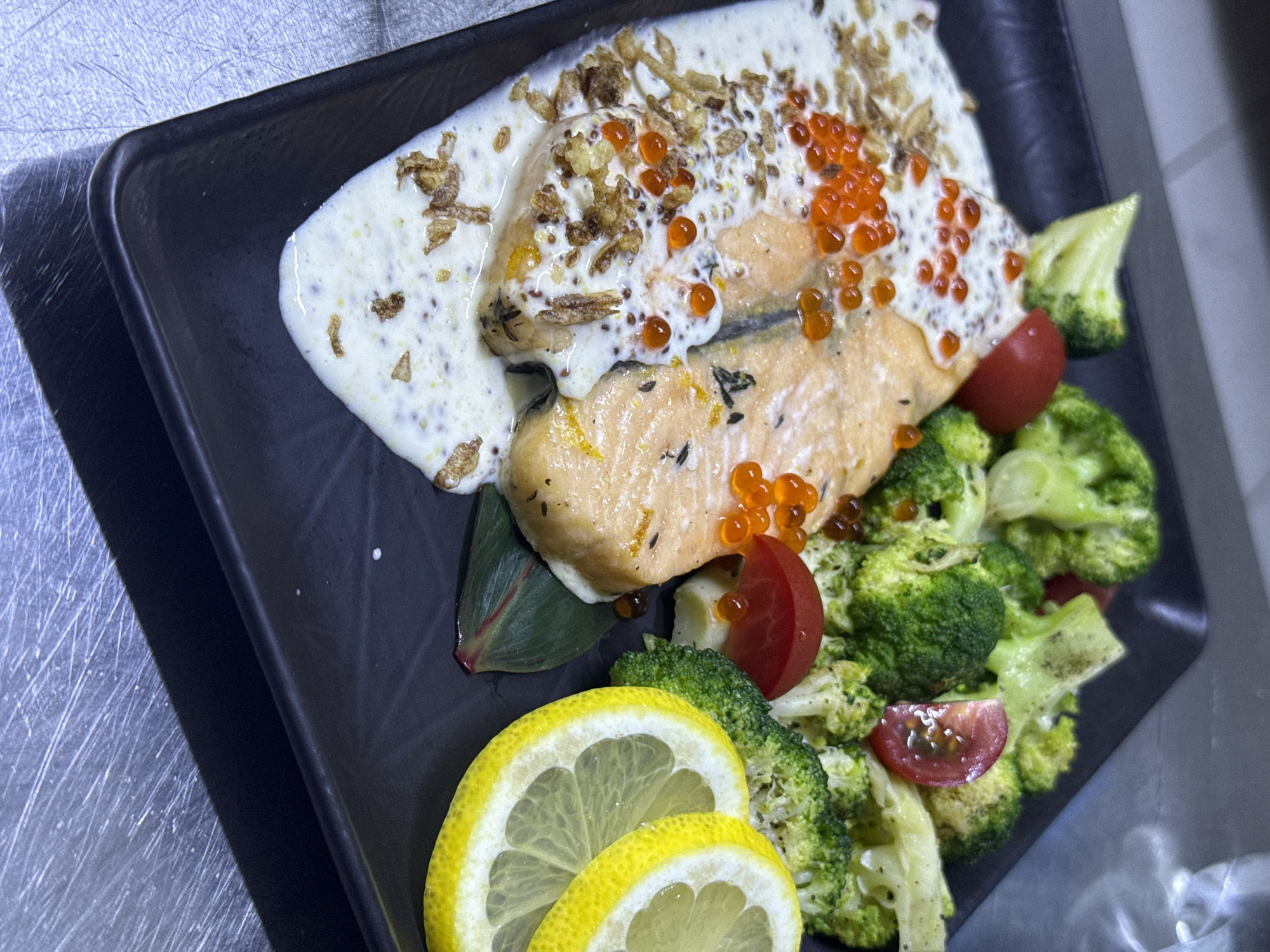 A close-up, overhead shot showcases a gourmet dish of salmon served on a black square plate. The salmon fillet is bathed in a creamy white sauce, garnished with bright orange salmon roe and crispy fried onions. Accompanying the fish are steamed broccoli florets, halved cherry tomatoes, and two slices of lemon. The dish is presented on a textured metallic surface. The image provides no information about the location in the city of Ақши, Kazakhstan.