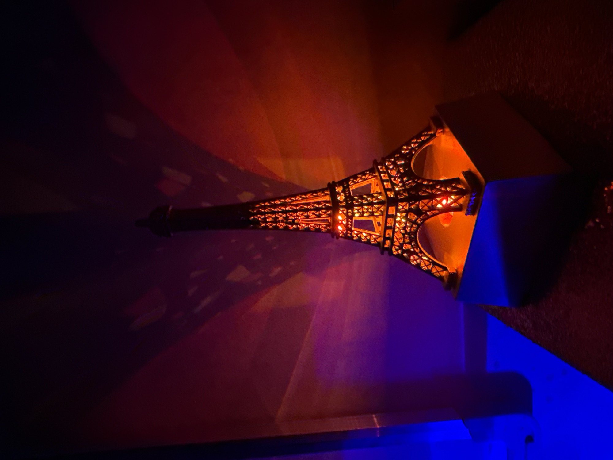 An illuminated replica of the Eiffel Tower stands on a square, light-colored pedestal. The replica is made of dark, intricate metalwork and is lit from within by a warm orange glow. The background is a wall with a gradient of colors, transitioning from deep purple on the left to a bright reddish-orange on the right. Shadows of the Eiffel Tower replica are cast onto the wall, creating a textured, abstract pattern. The lighting is dramatic, with strong contrasts between the illuminated tower and the darker areas of the scene. There are no people or animals visible. The scene is likely indoors, given the controlled lighting and the nature of the object. The location in the city of Opoczno, Poland is not determinable from the image itself.