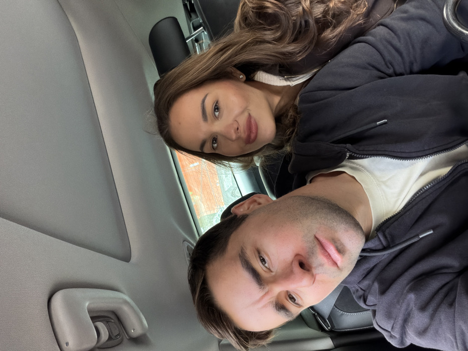 A self-portrait is captured inside a vehicle, depicting two individuals in Tel-Aviv, Israel. In the foreground, a male with short dark hair and faint stubble wears a black zipped hooded sweatshirt over a light-colored t-shirt, looking towards the camera with a neutral expression and slightly furrowed brows. Behind and to his right, a female with long wavy brown hair wears a dark zipped jacket with a light-colored collar, looking at the camera with a subtle smile. A small stud earring is visible in her right ear. The vehicle's interior features a grey headliner, a functional grab handle on the upper left, and dark-colored seats with visible headrests. Through the rear window, a reddish-brown brick wall is clearly visible in the outdoor background, suggesting the vehicle is stationary or moving slowly alongside a building.
