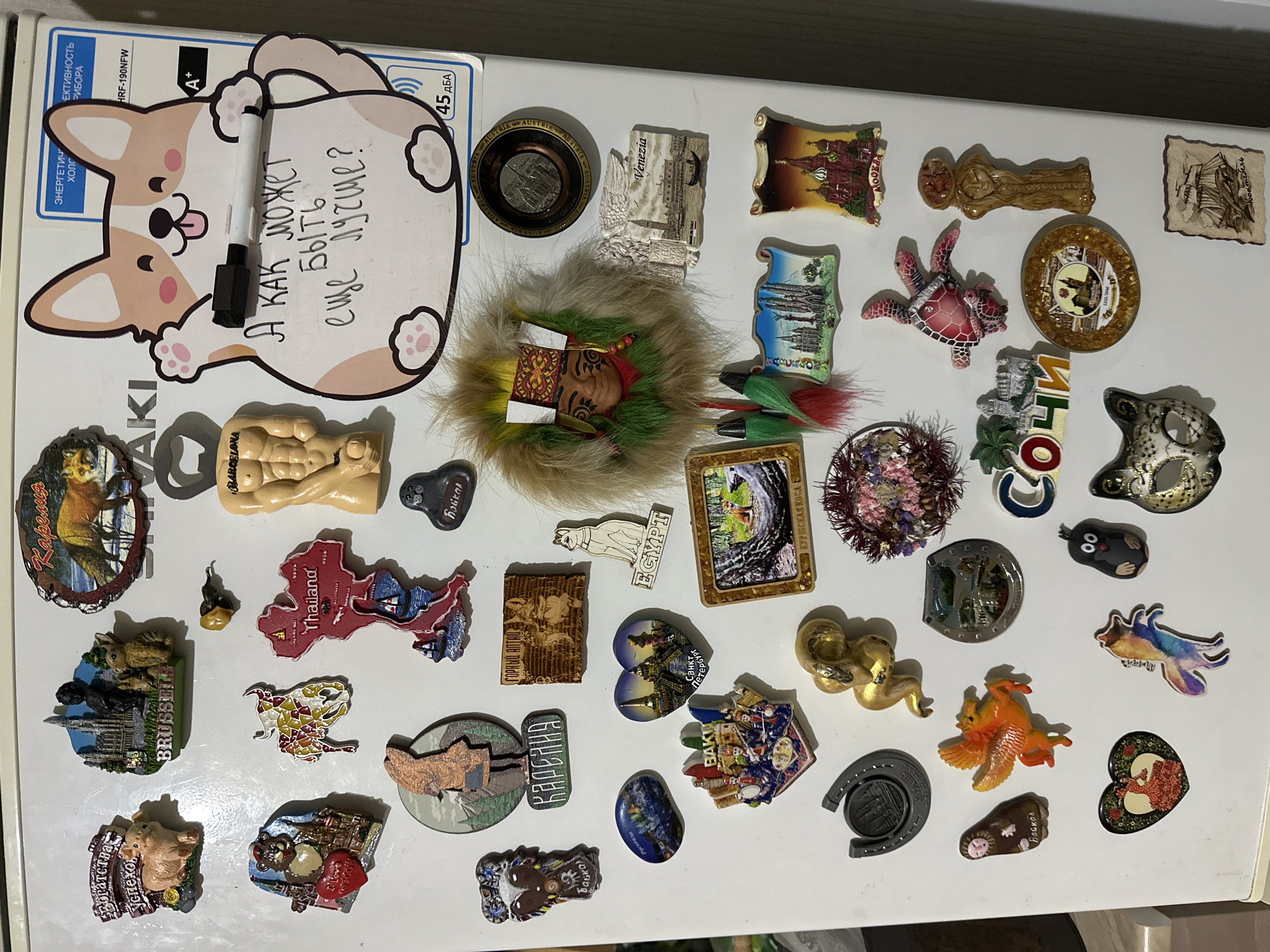 The image shows a refrigerator door covered in magnets from various places around the world. There are recognizable destinations like Barcelona, Thailand, Brussels, Venice, Egypt, and Sochi. Some magnets are shaped like landmarks, while others feature animals or decorative designs. There is a dry erase board in the shape of a dog with the phrase "А как может быть еще лучше?" written in Russian, which translates to "How can it be even better?" A dry erase marker is stuck to the board.