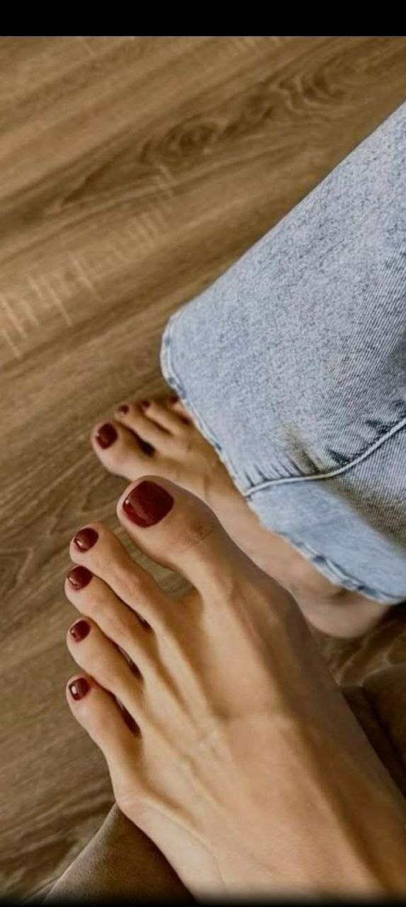 The image shows a pair of bare feet with dark red painted toenails resting on a light brown wooden floor. The legs are clad in light blue jeans. The location is not specified, and there is no indication of the city of Minsk, Belarus.