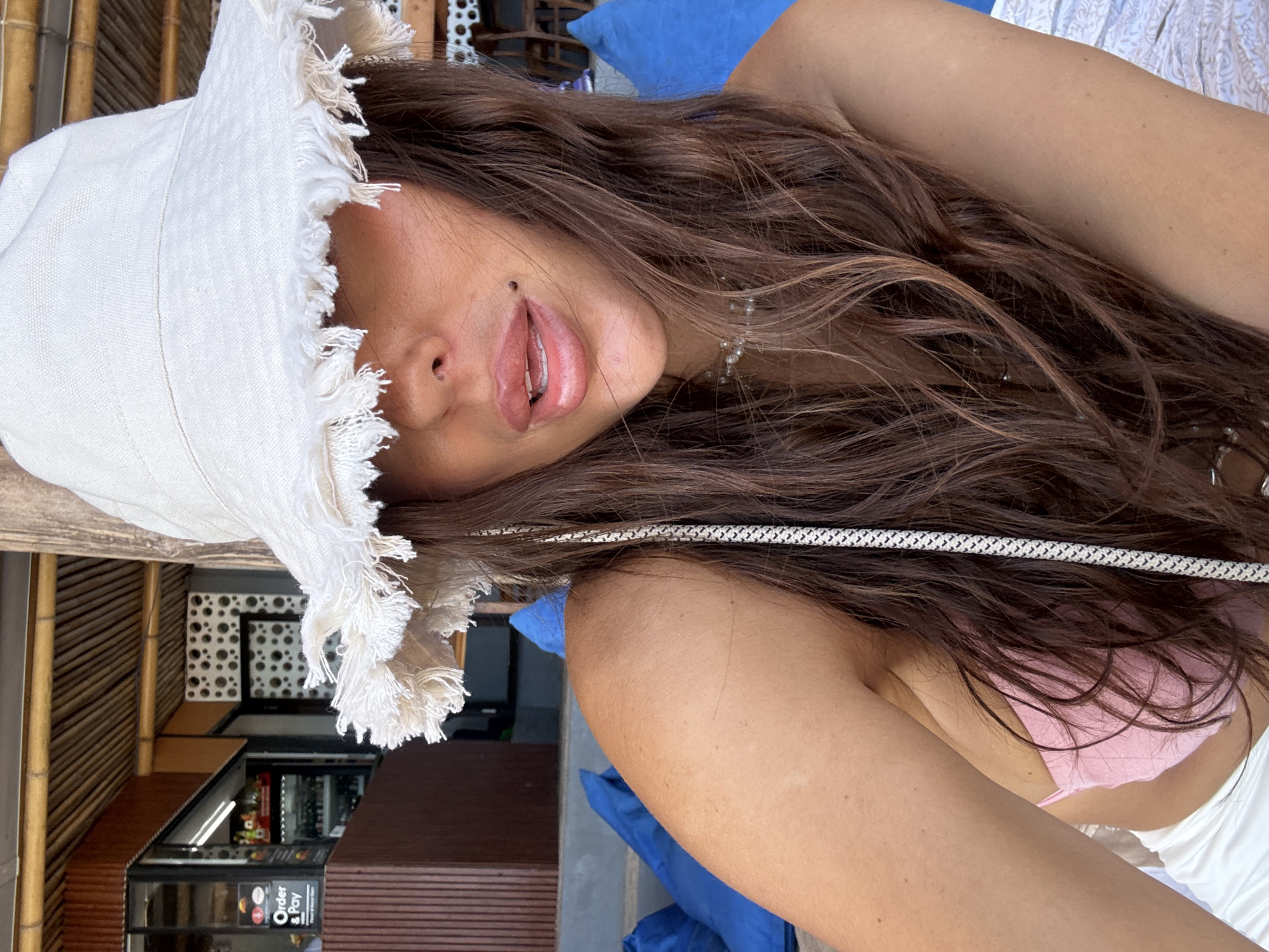 A woman with long brown hair is reclining outdoors, likely at a restaurant or cafe. She is wearing a white bucket hat with a fringed brim and a pink bikini top with a white cover-up. The location appears to be Kuta Selatan, Indonesia, as a sign reading "Order & Pay" is visible in the background. The overall composition of the image suggests a relaxed, casual setting.