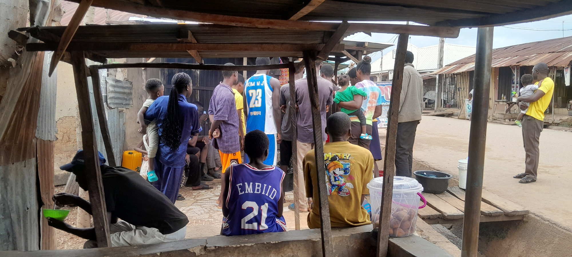 The image depicts an outdoor market scene, likely in Abuja, Nigeria, under a makeshift shelter constructed with corrugated metal roofing and wooden supports. Several individuals are present, appearing to be a mix of adults and children. In the foreground, a person wearing a blue baseball cap and black t-shirt is crouched, tending to a green bowl with a ladle. To their right, another person is seated, wearing a yellow t-shirt with a cartoon character graphic and the word "MICKEY" visible. Behind them, a group of people are standing. One individual wears a blue and white basketball jersey with the number 23 and text that appears to be Korean. Another person is holding a young child. A man in a yellow t-shirt holds a child and stands on the right side of the frame. The environment is dusty with a dirt road visible in the background, lined with simple buildings and structures, some with corrugated metal roofs. Wooden planks create a makeshift walkway over a trench or drainage area. Various containers, including buckets and a large yellow jerrycan, are scattered around the scene. The overall impression is one of a humble, community-based market setting.